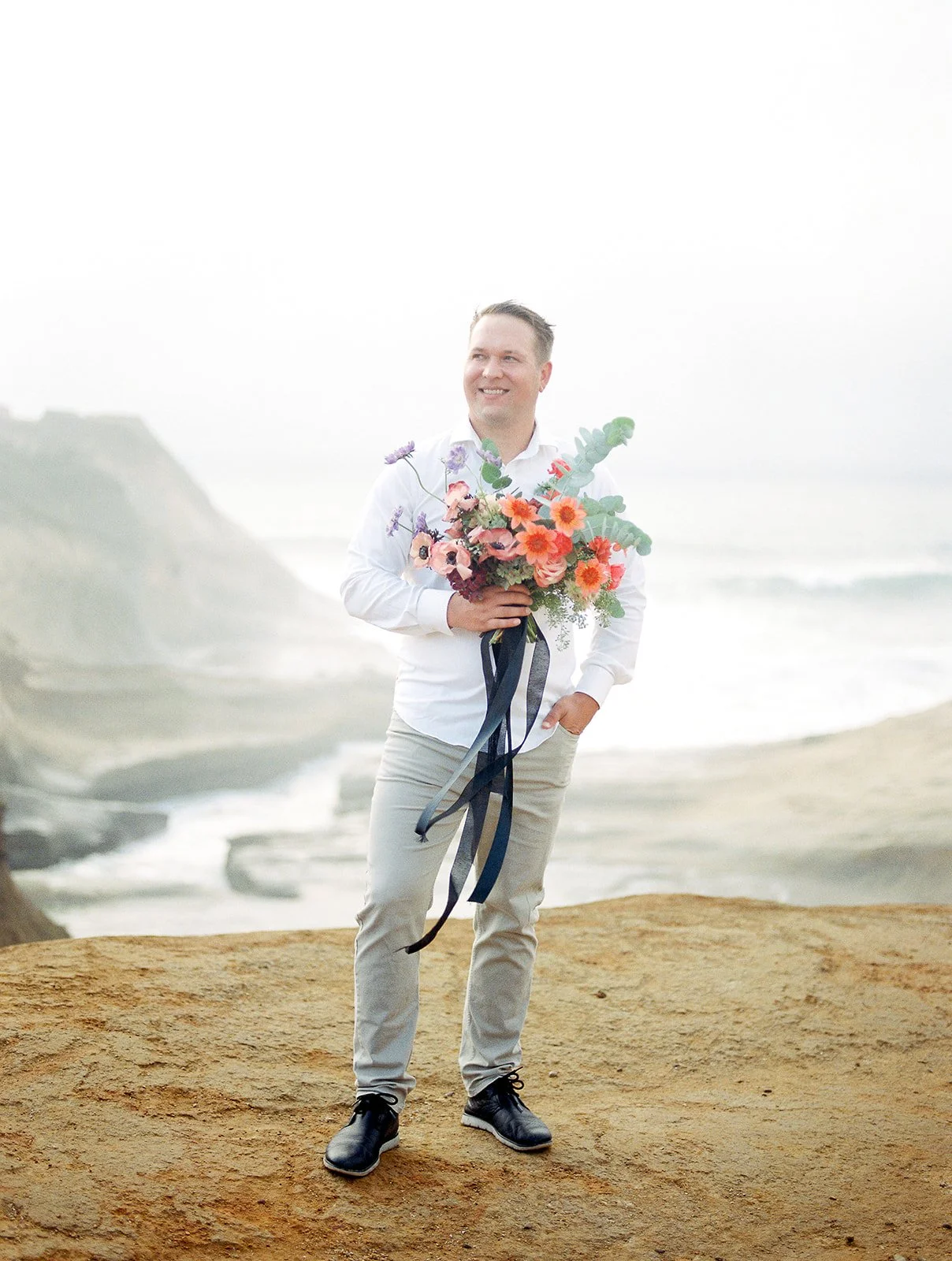 Man holding vibrant bouquet standing on coastal cliff with ocean and rock formations in background