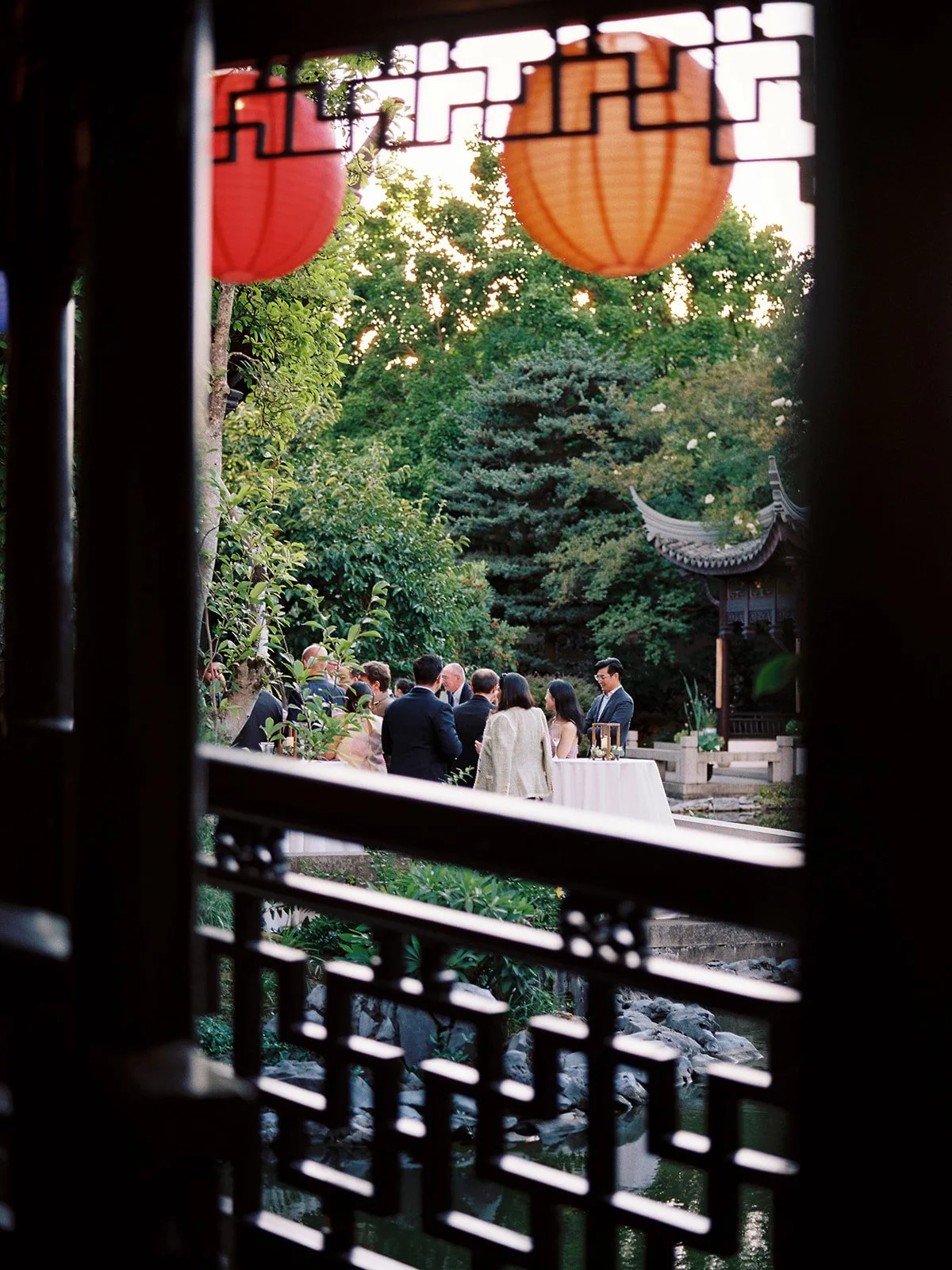 Evening view through a latticed window frame with red and orange paper lanterns, looking out to guests gathered around a candlelit table in the garden courtyard.