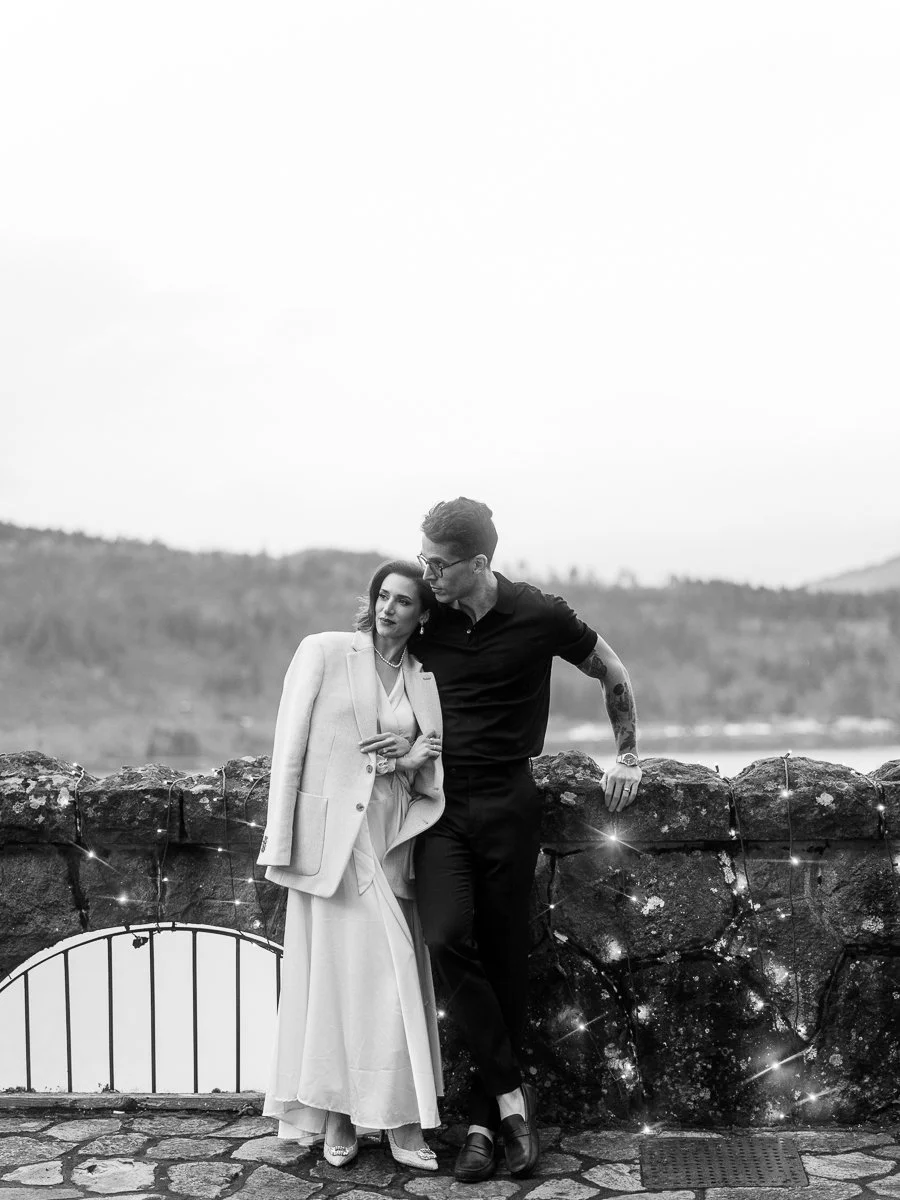 Black and white portrait of a couple leaning against a stone wall strung with fairy lights, river and hills behind them.