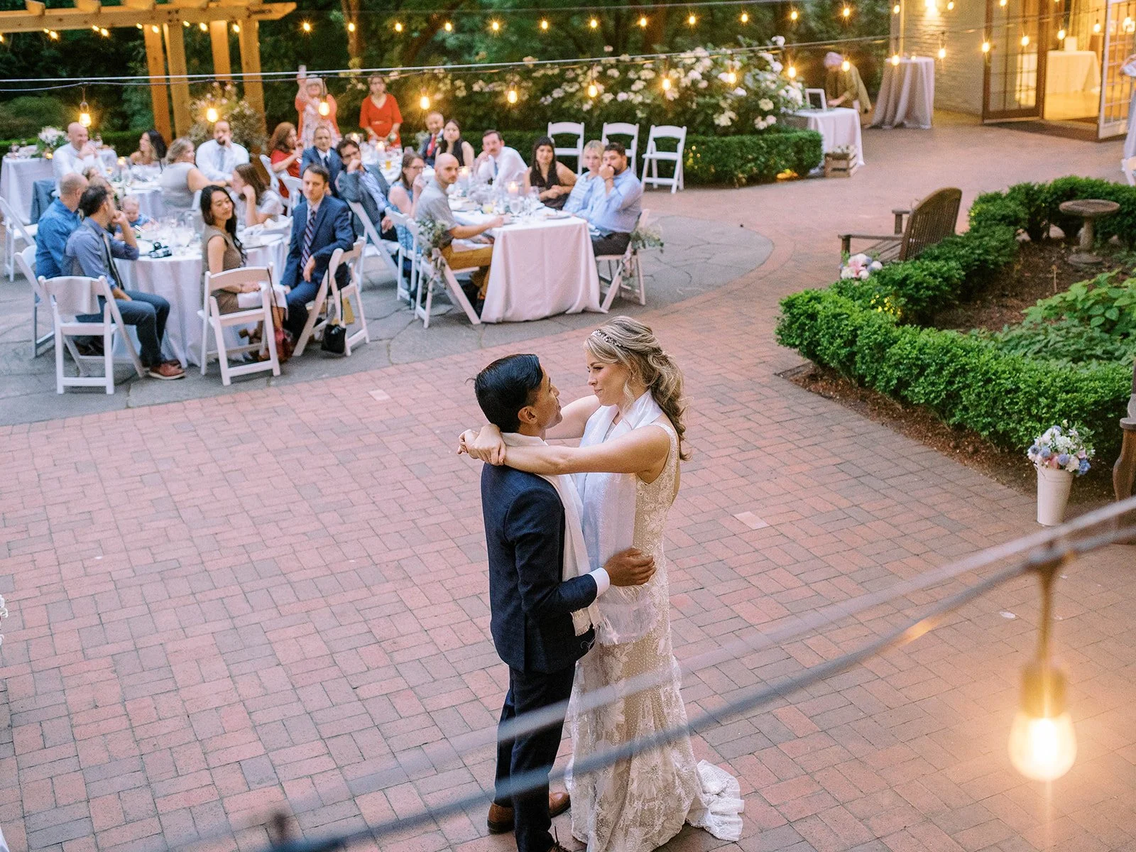 Newlyweds slow dance on the brick patio while guests watch from their reception tables.