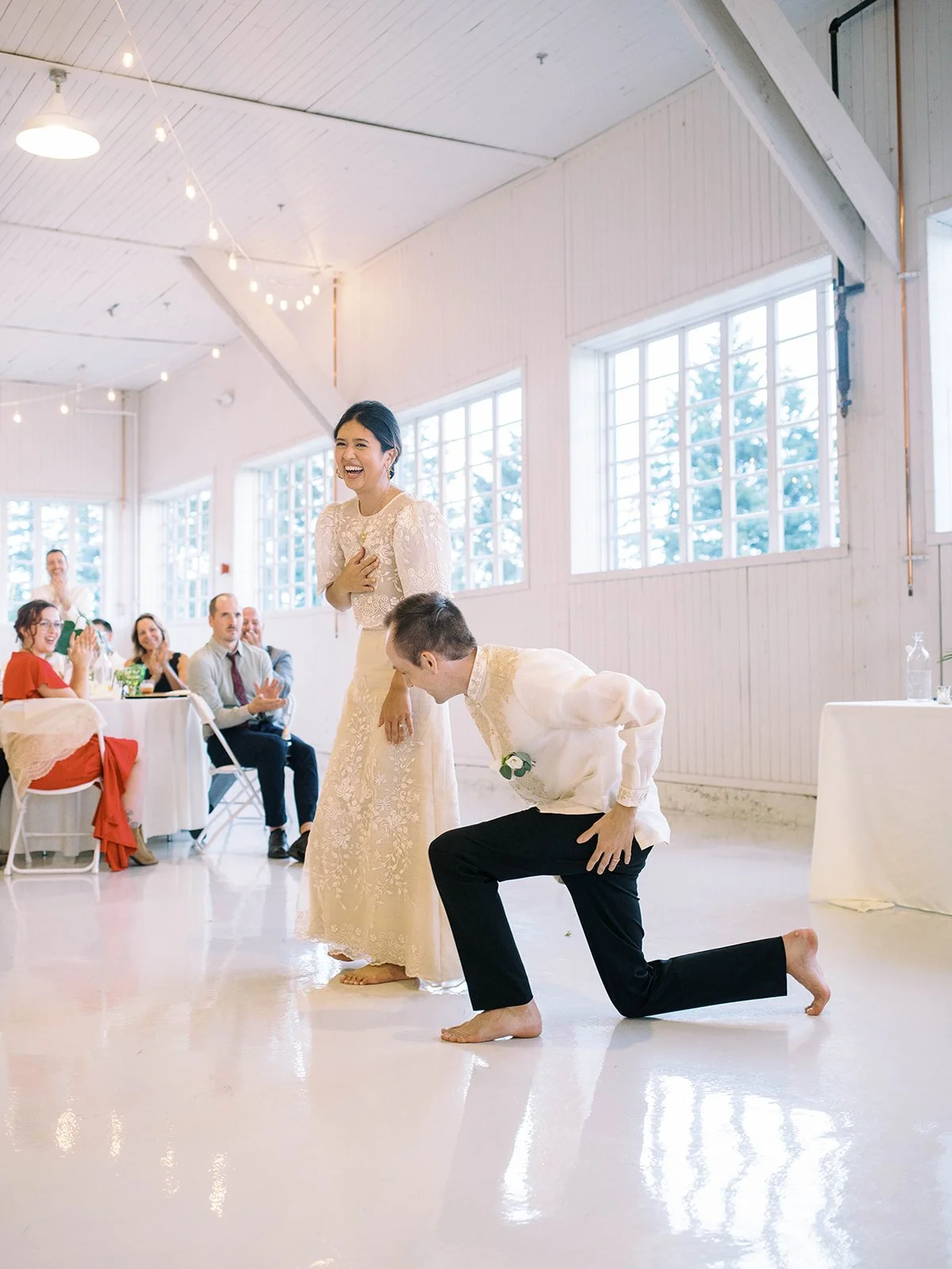 Groom taking a dramatic barefoot bow on the hangar dance floor while the bride laughs with guests applauding in the background