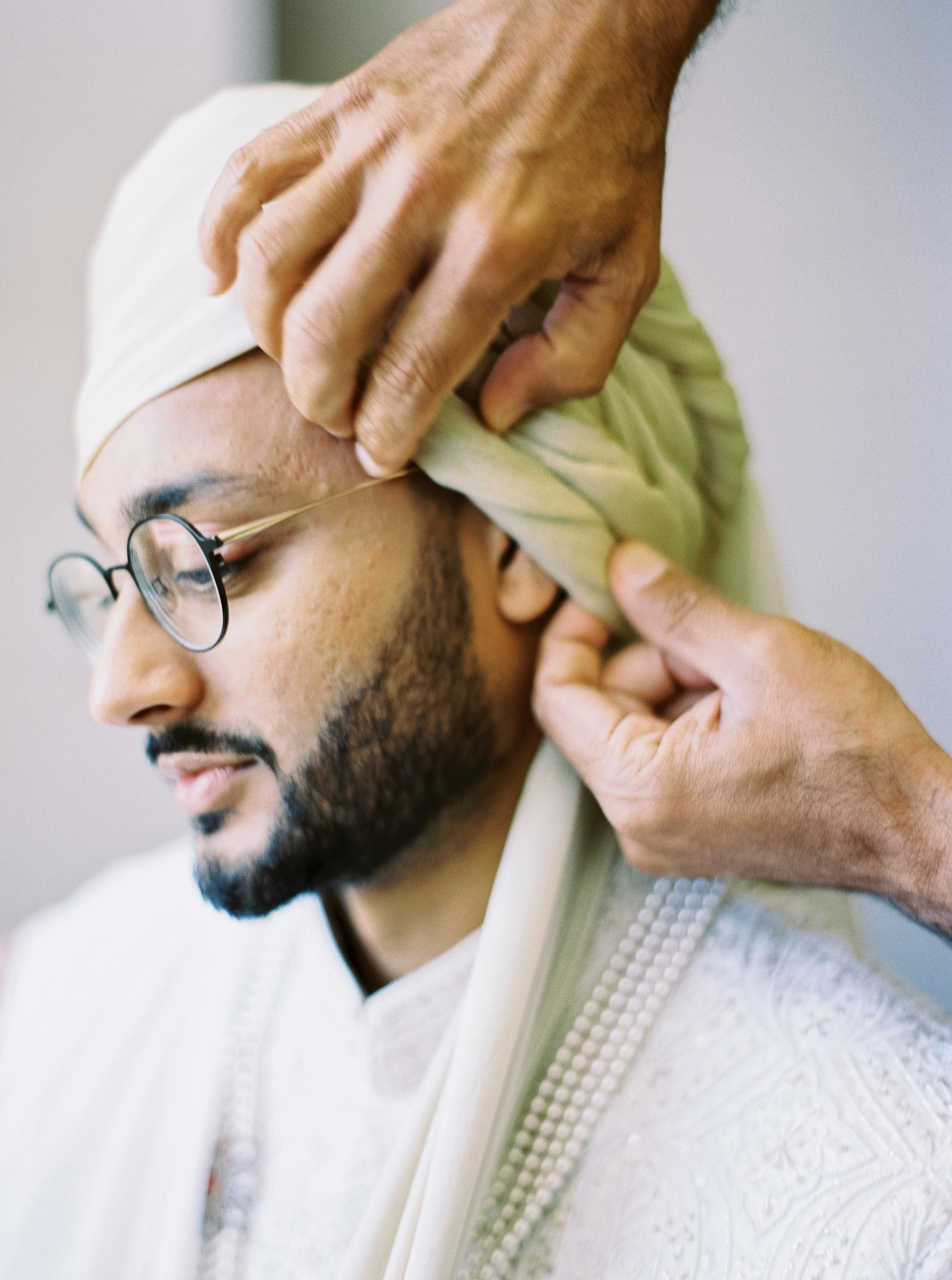Hands carefully wrap a pale green turban around a groom wearing embroidered white wedding attire.