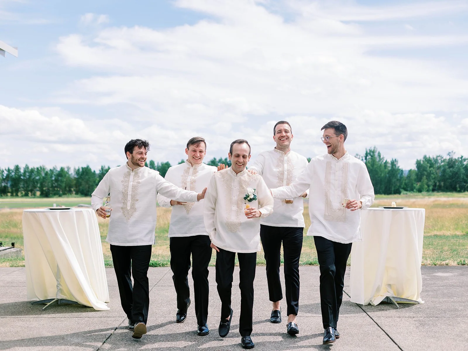 Five groomsmen laughing and walking together in matching white embroidered barong tagalog shirts and black trousers at an airfield
