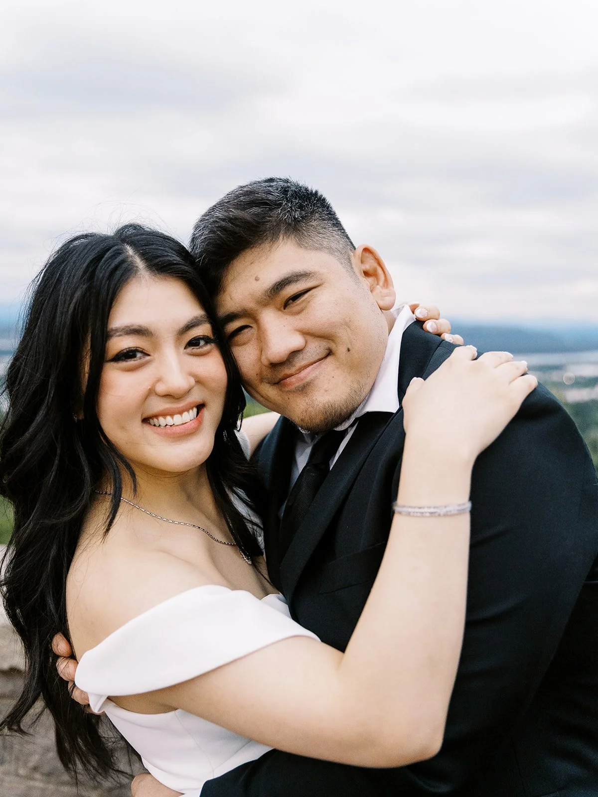 Close portrait of the bride and groom embracing and smiling at the camera at The Grotto overlook, the cloudy Portland skyline stretching out behind them