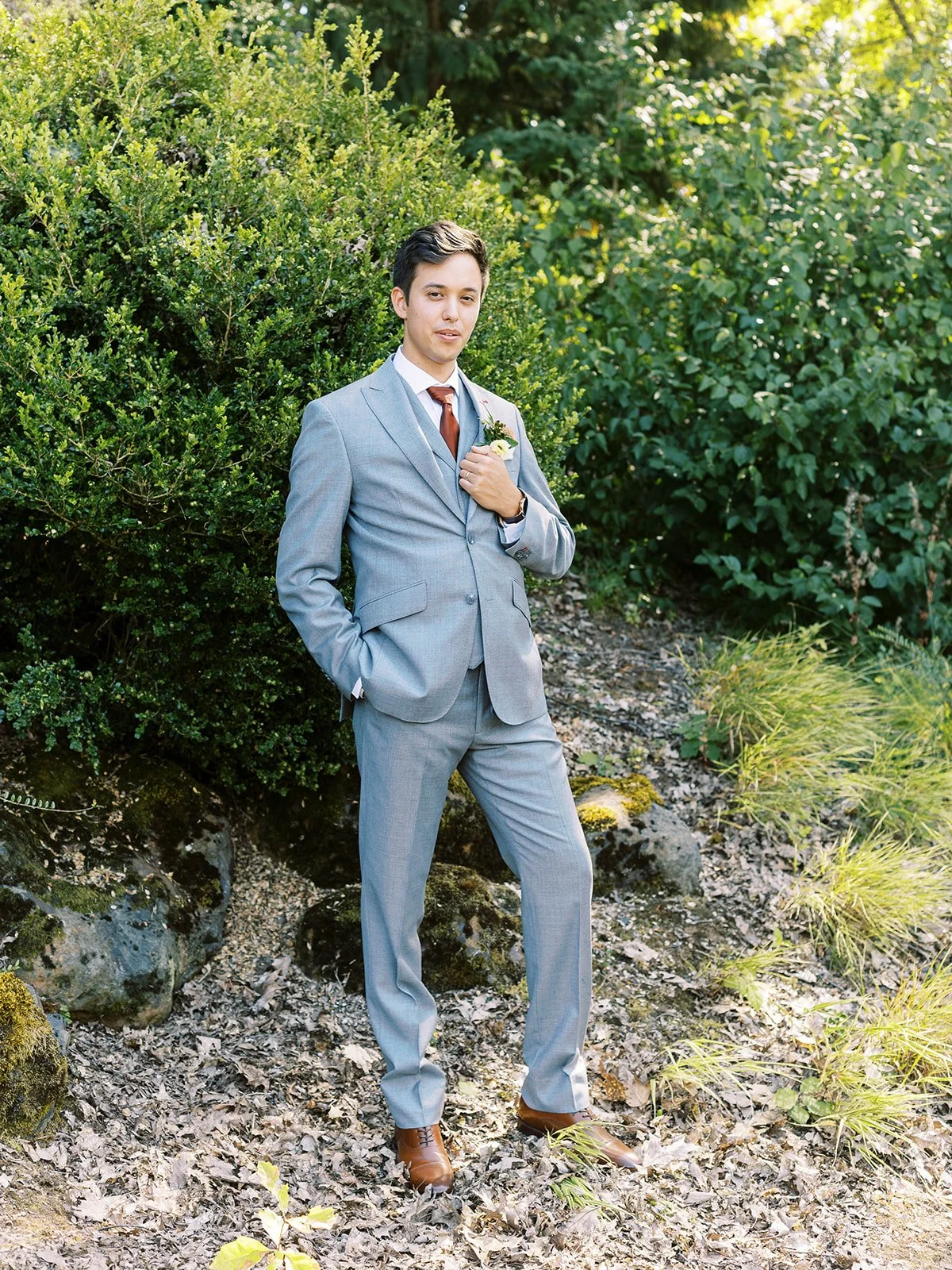 Groom standing outdoors in a light gray suit with a rust tie and floral boutonniere.