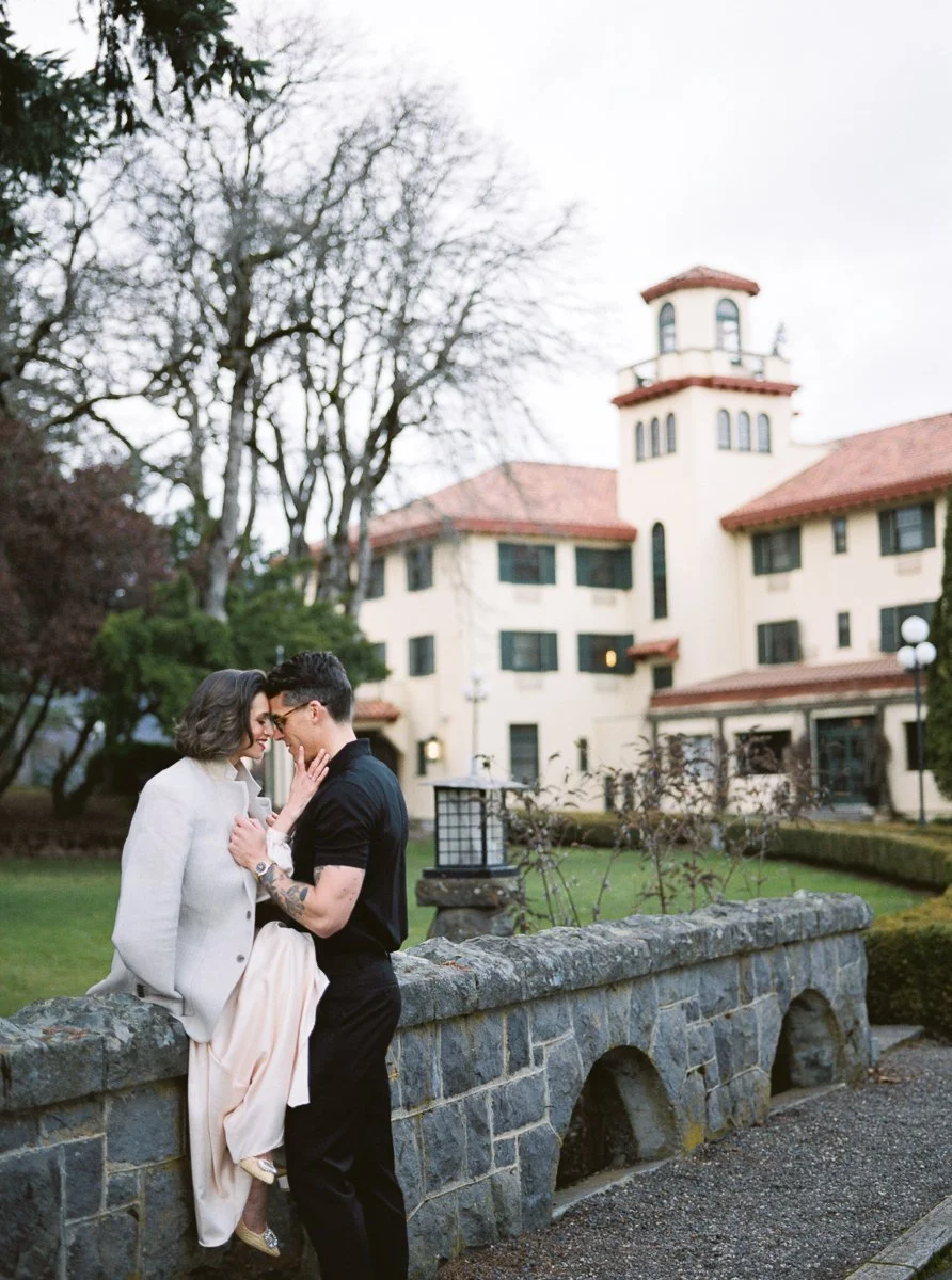 Couple standing close by stone wall, leaning in forehead to forehead outside historic building with trees and hedges