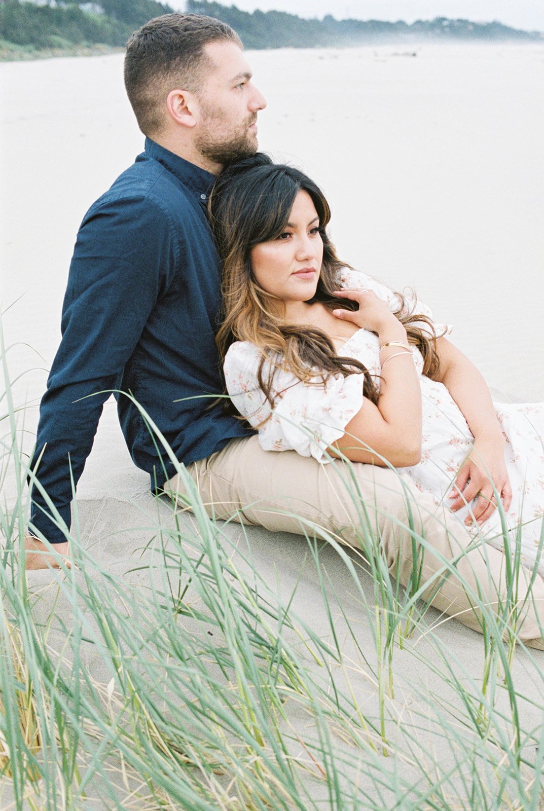 Couple seated in beach grass, woman leaning back against man with soft coastal background