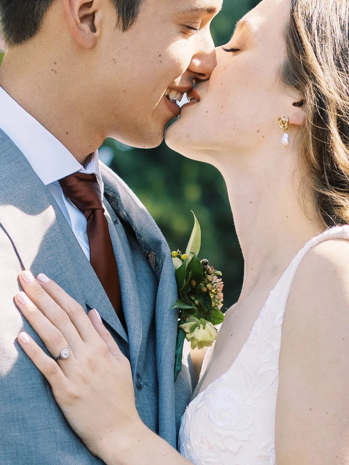 Close-up of bride and groom kissing, showing wedding ring and boutonniere in natural outdoor light.