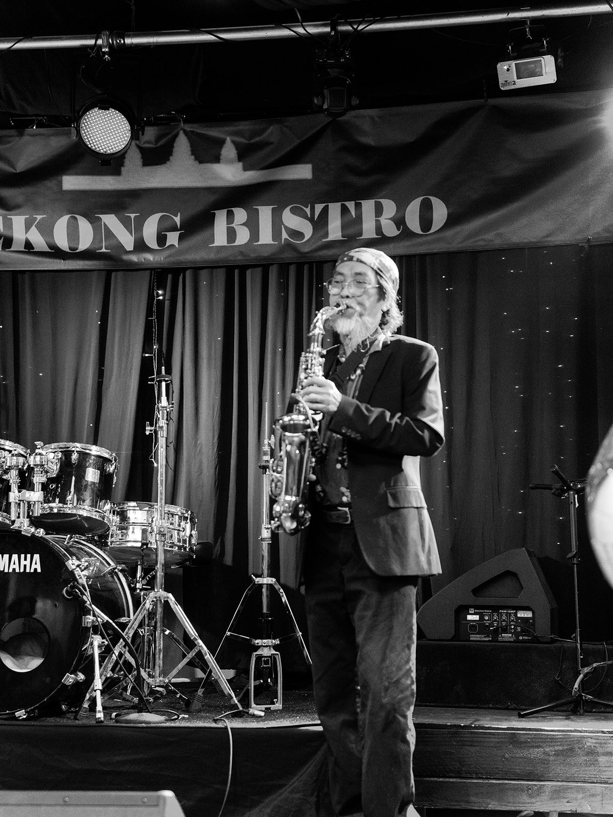 Black and white photo of an older saxophonist with a white beard performing on the Mekong Bistro stage, a Yamaha drum kit behind him