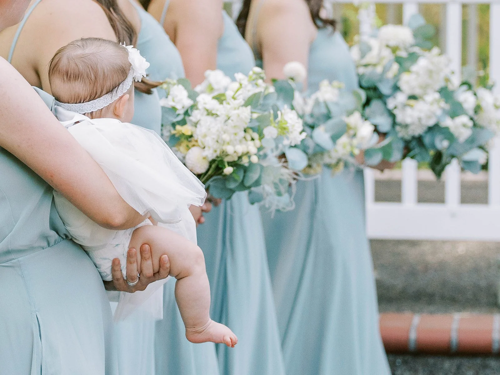 Bridesmaid holding baby flower girl in white dress and floral headband during garden wedding ceremony.