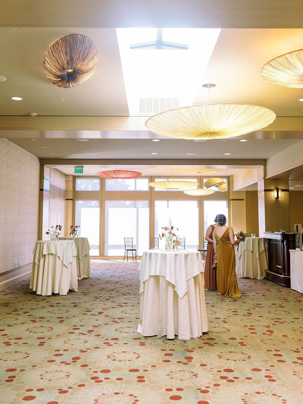 Bright hotel cocktail hour space with large disc pendant lights and a skylight, featuring white cocktail tables with small floral centerpieces and two bridesmaids in gold and rust dresses