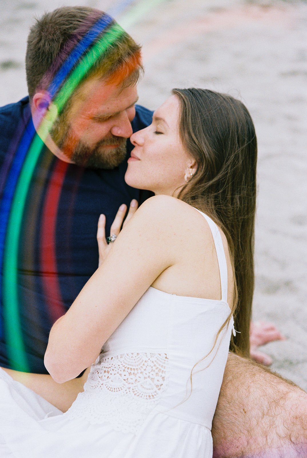 Couple sitting on sand, leaning into each other with a subtle rainbow light leak across the frame.