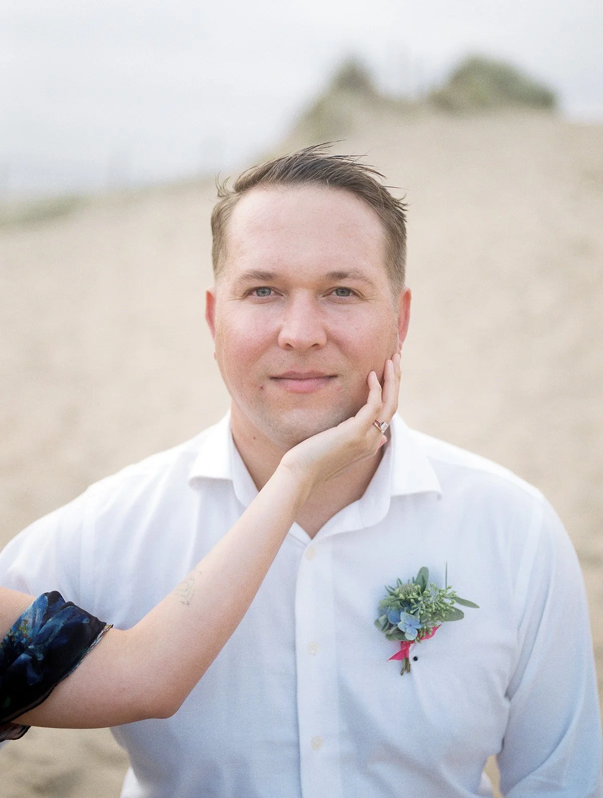 Portrait of groom with boutonniere as partner’s hand rests on his cheek, soft coastal backdrop