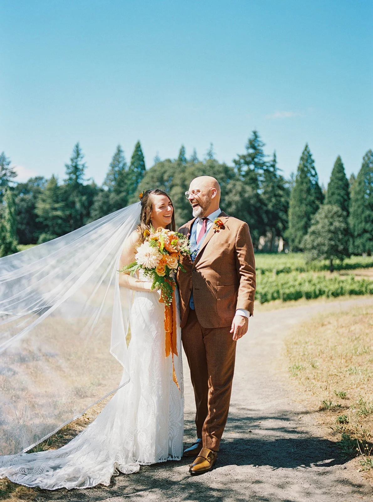 Bride and groom walking together, smiling, veil flowing in outdoor vineyard setting