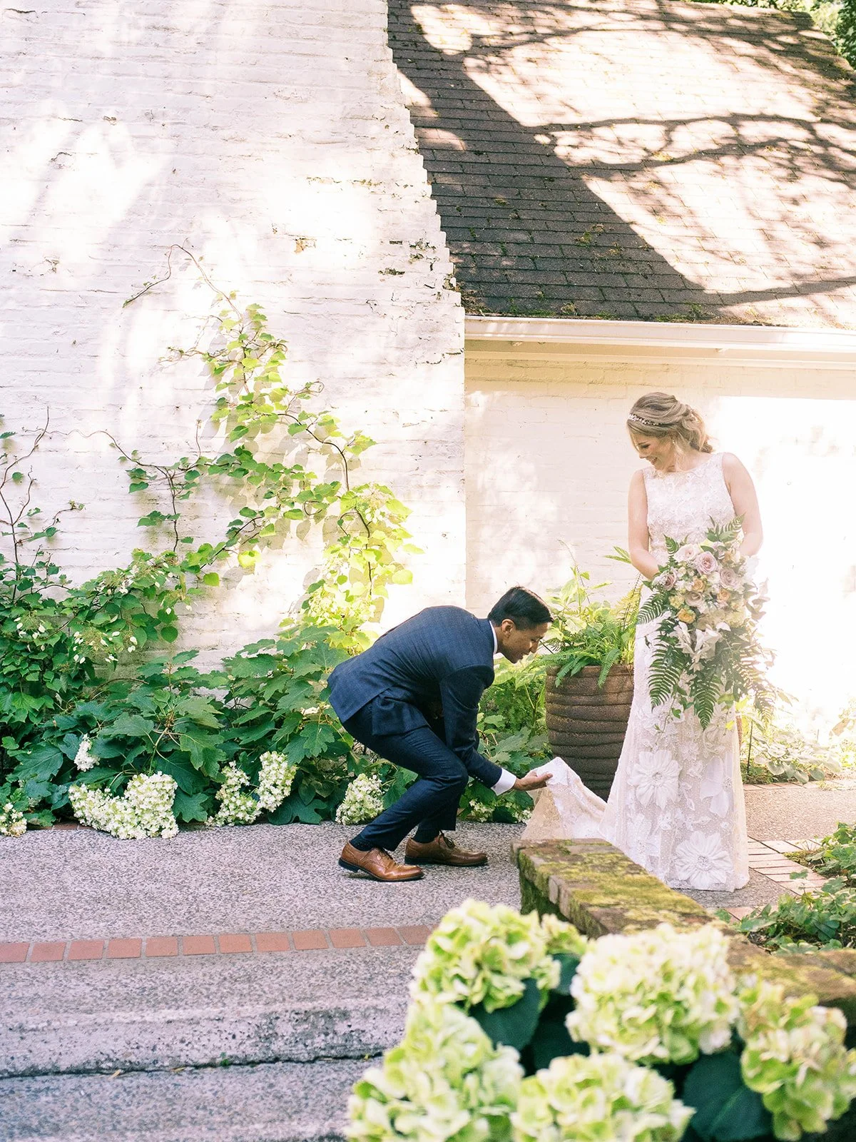 Groom bends to adjust the bride’s dress as she holds a cascading floral bouquet.