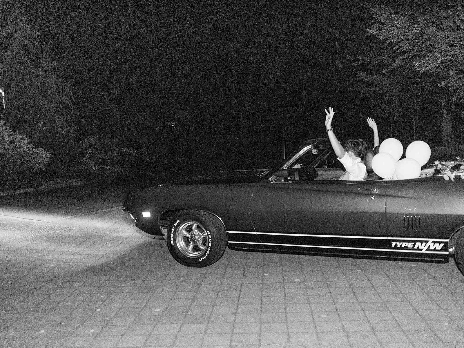 Black-and-white photo of the newlyweds driving away in a vintage convertible with balloons in the back.