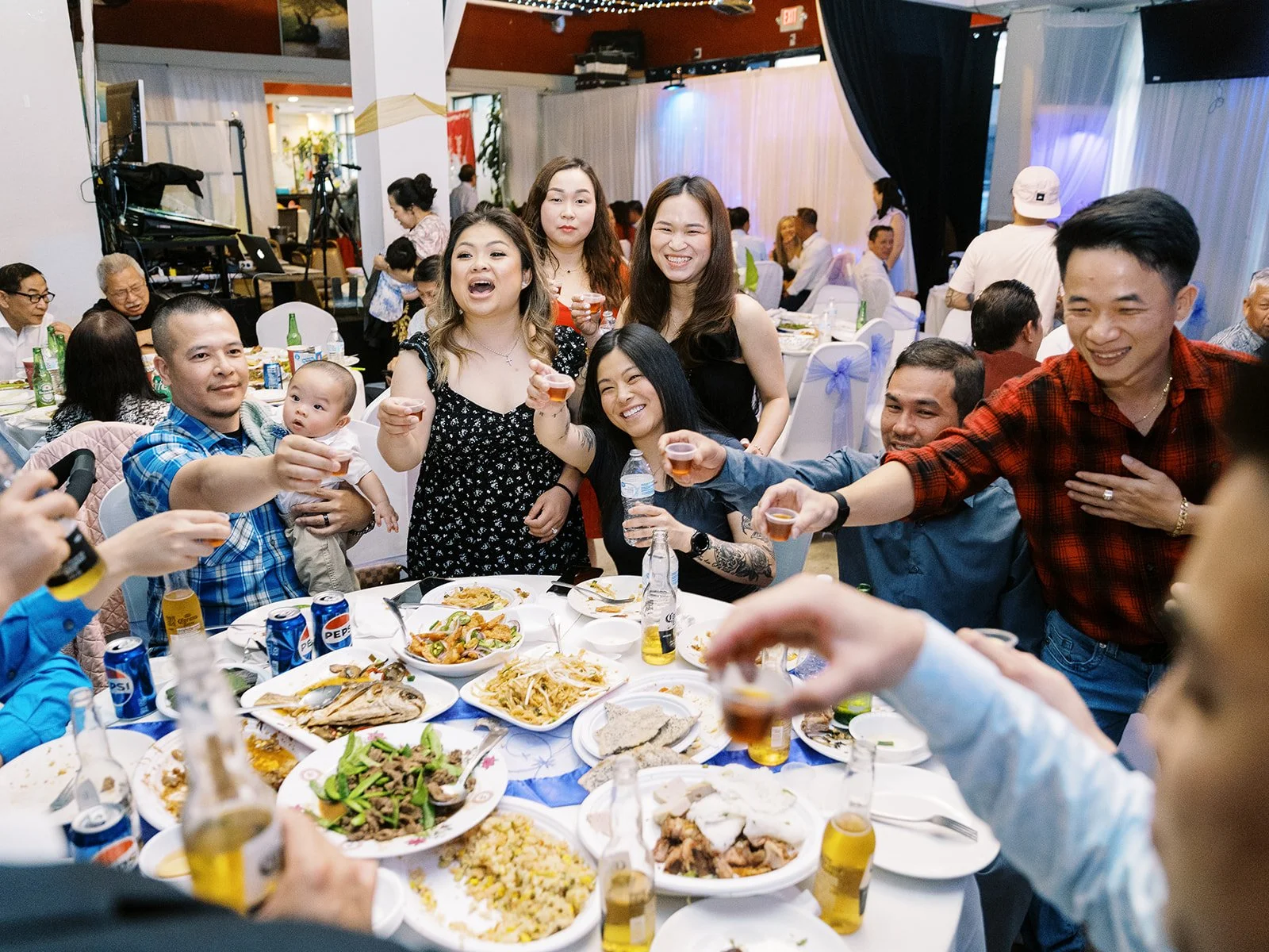 Energetic table toast at the wedding reception, guests reaching in with shot glasses over a spread of shared dishes, a baby held by one of the guests