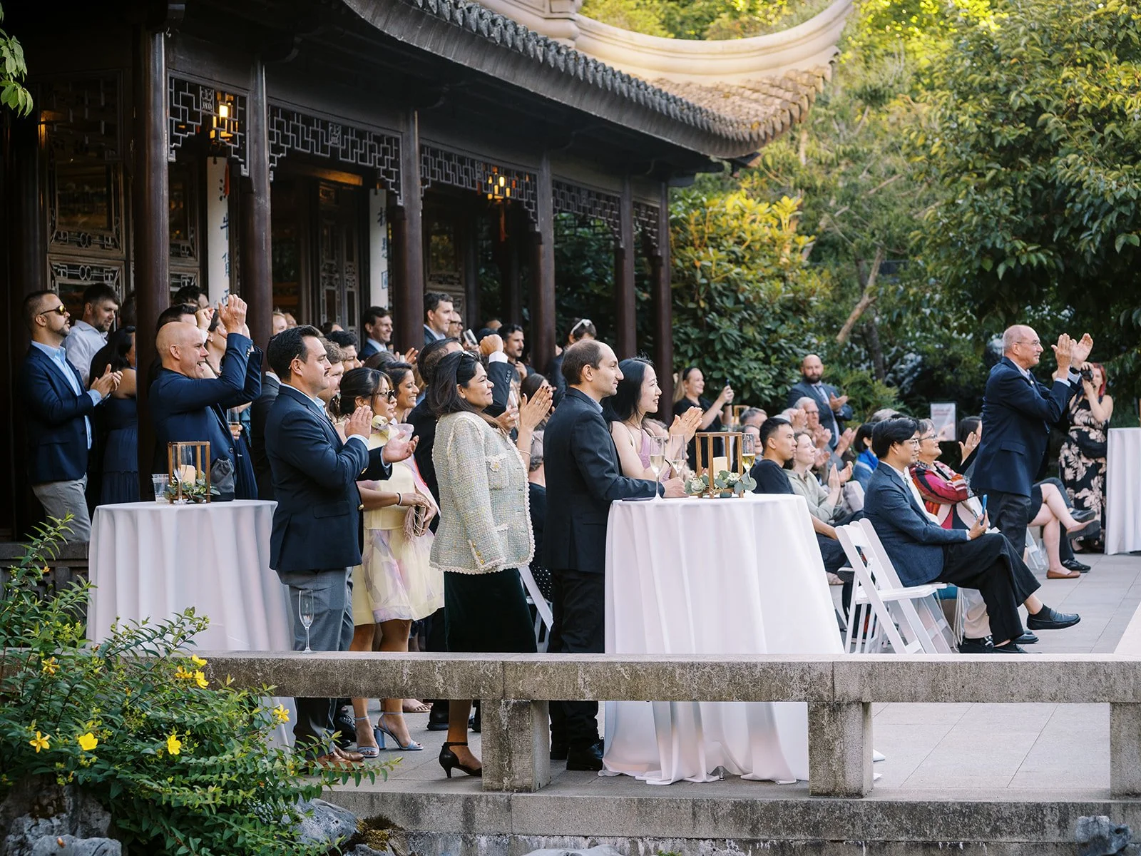 A large crowd of wedding guests applaud along the garden terrace, standing and seated at white-clothed cocktail tables beneath the dark wood pavilion eaves.