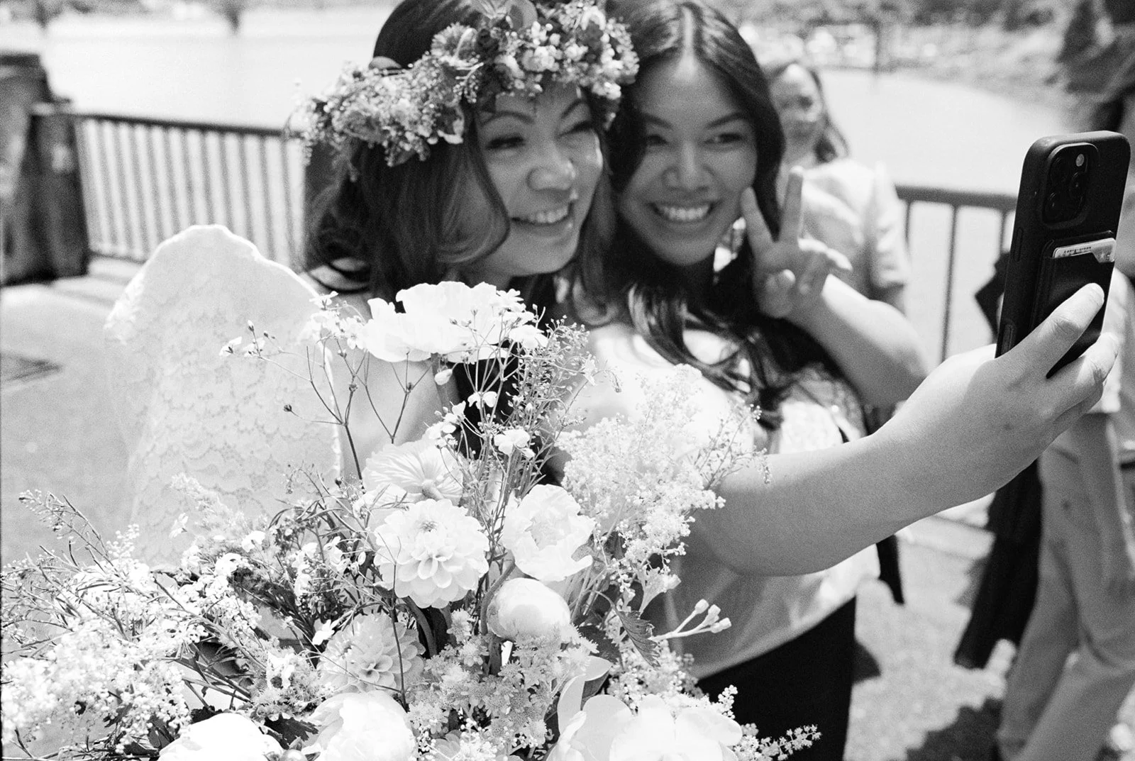 Black and white close-up of a smiling bride and wedding guest taking a selfie beside a large floral bouquet.