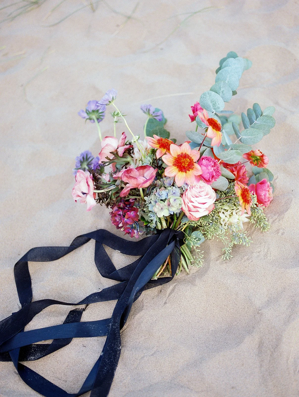 Colorful bridal bouquet with black ribbon resting on sandy beach, soft natural light