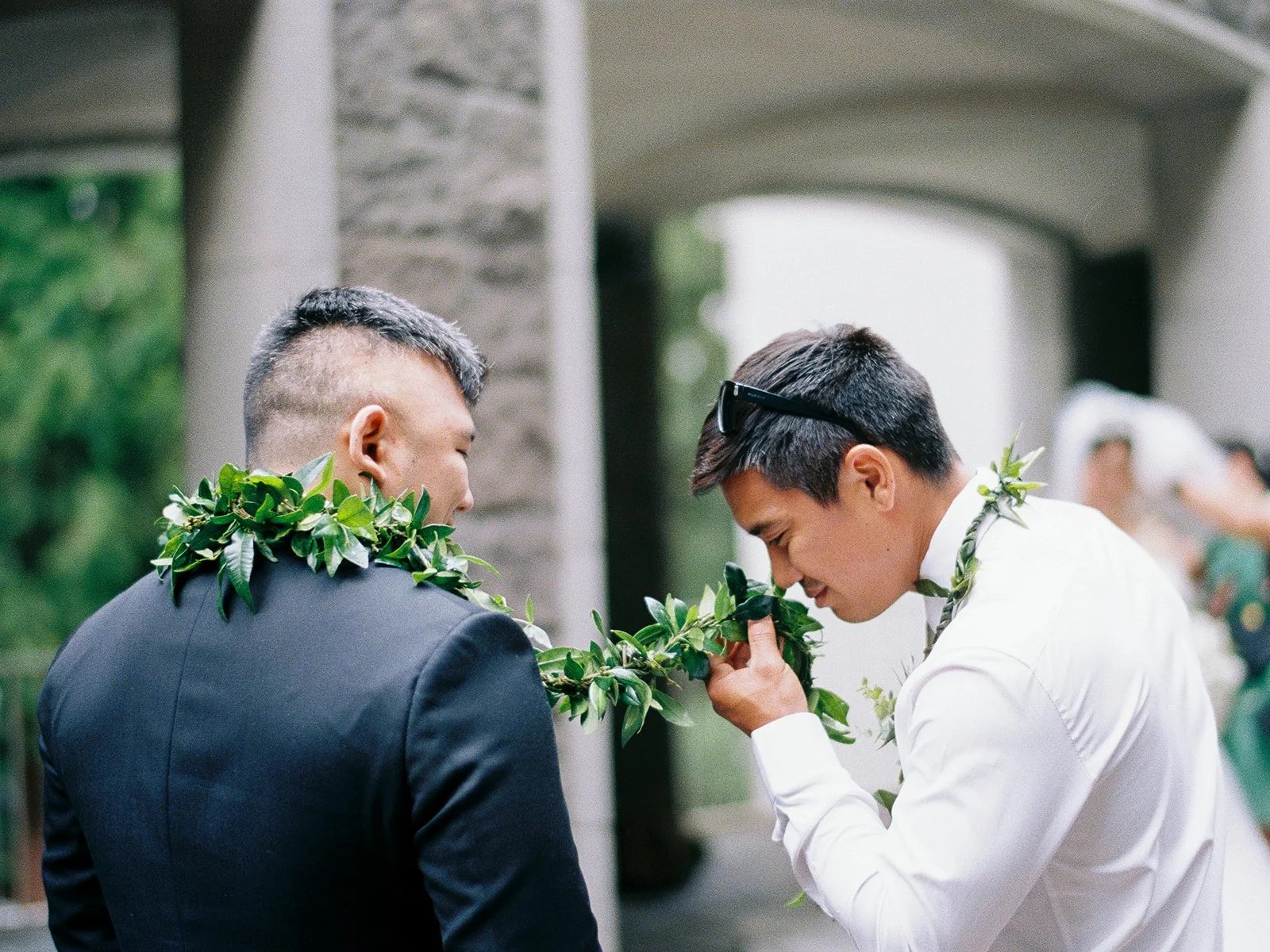 Two men in suits wearing green maile lei lean in close, one smelling the lei, with the bride and wedding party blurred in the background outside The Grotto chapel