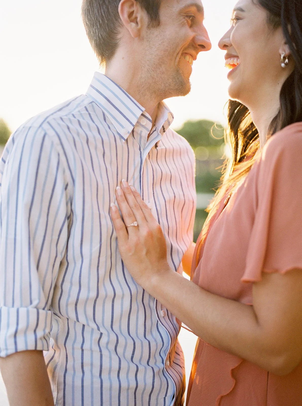 Close-up of couple smiling face to face at sunset, woman’s hand with engagement ring resting on partner’s striped shirt in warm light