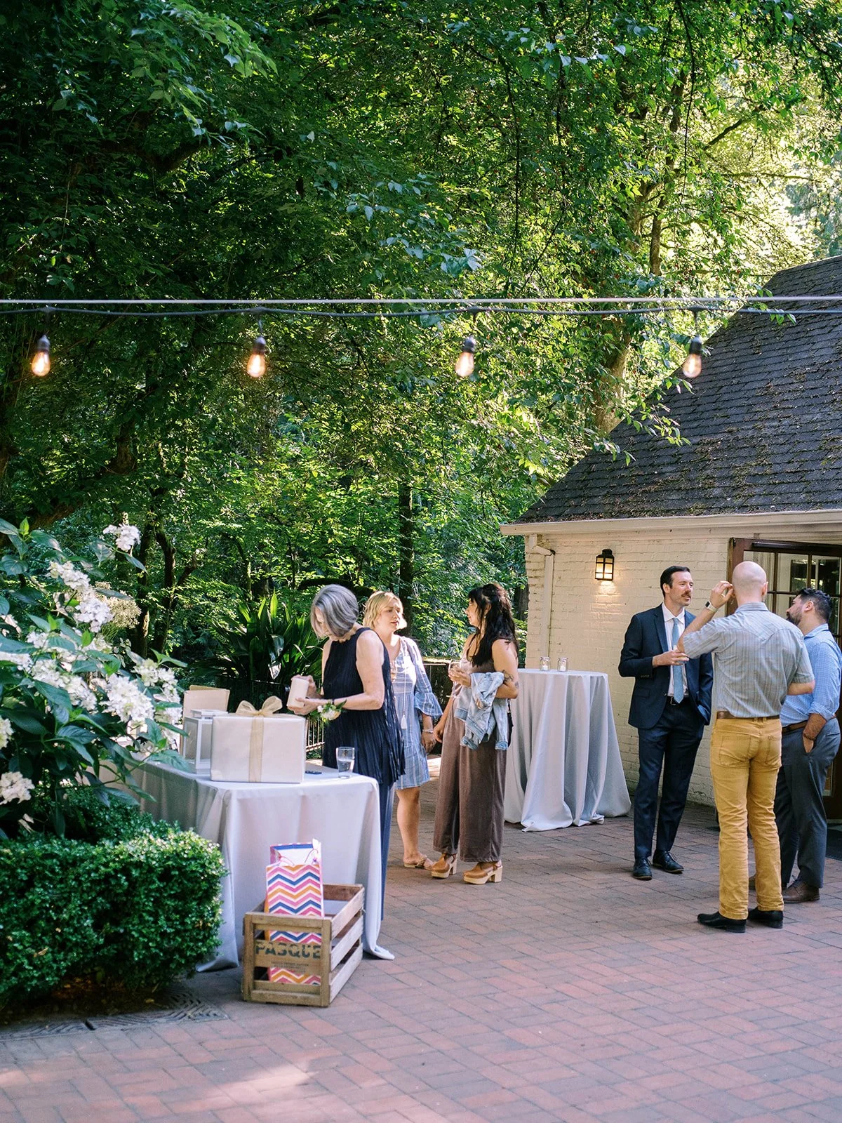 Wedding guests mingle beneath string lights on a garden patio surrounded by trees.