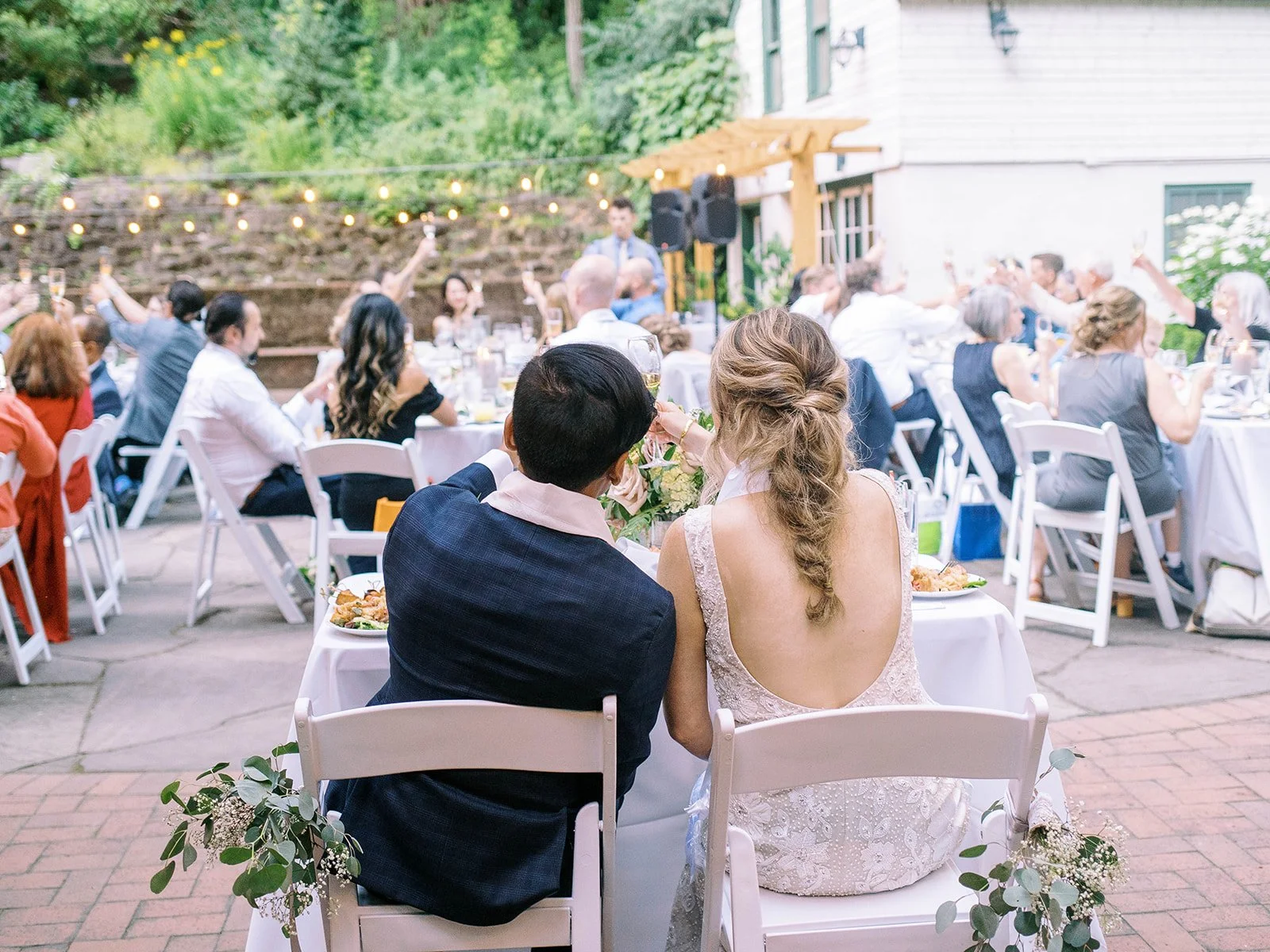 Bride and groom sit together at their reception table while guests raise glasses behind them.