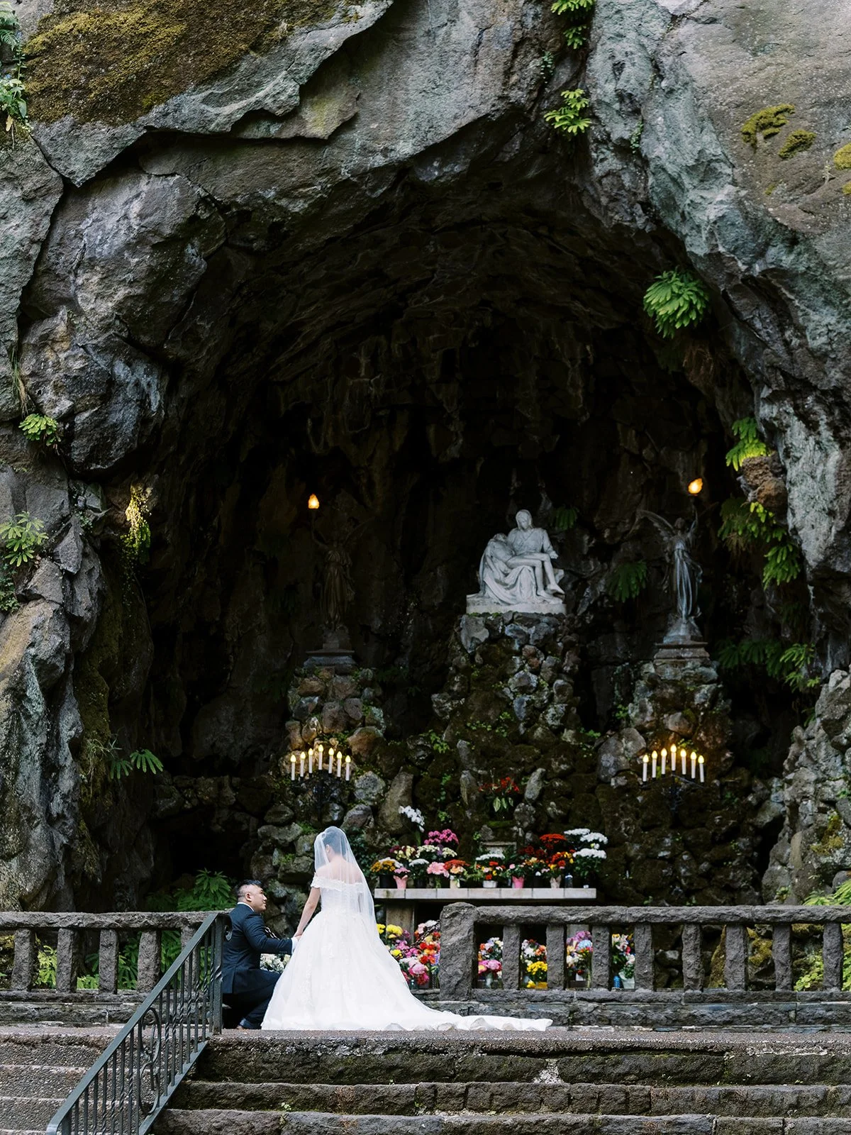 Bride and groom standing together before a grotto shrine carved into rock with candles and flowers.