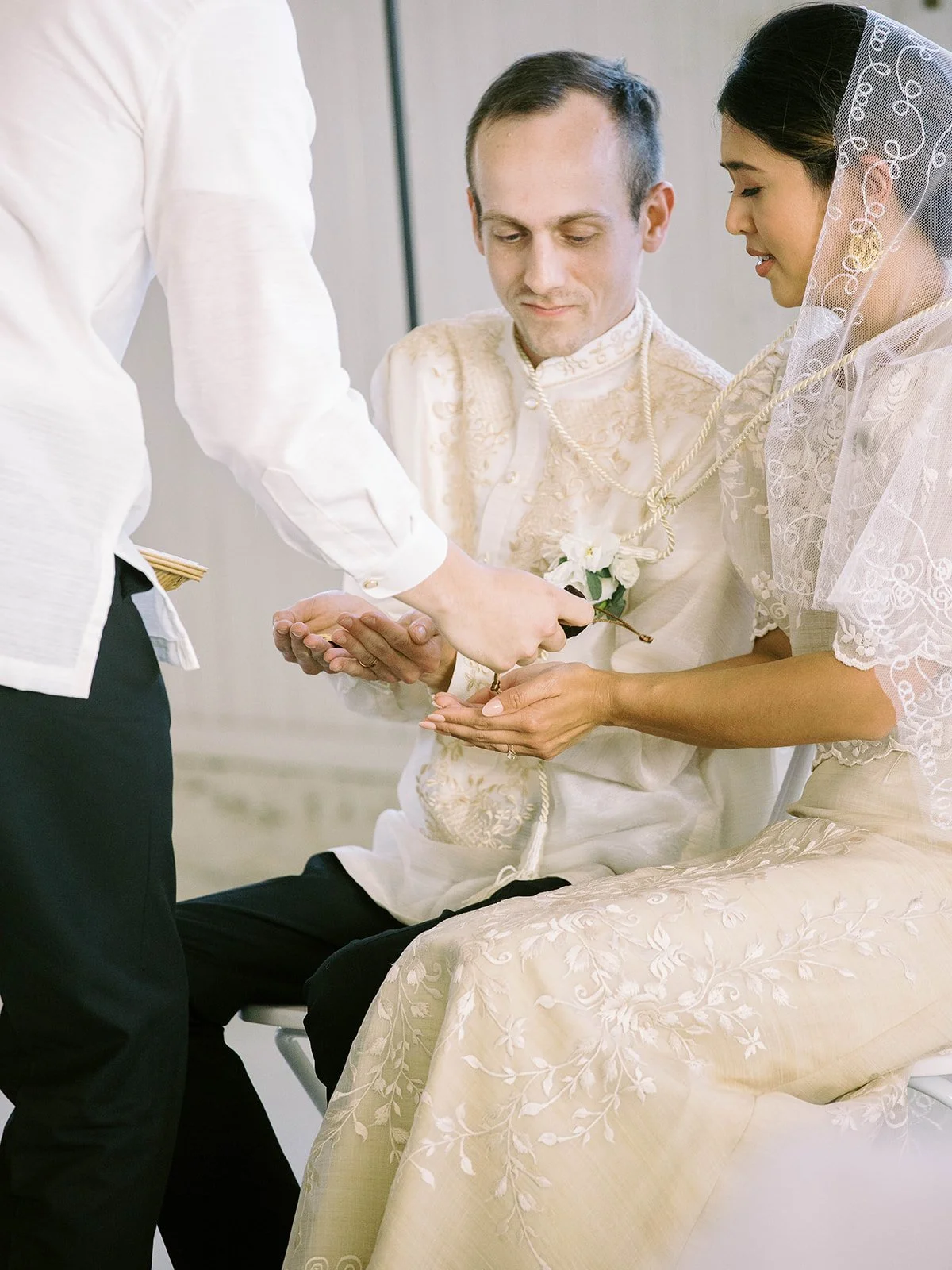 Close-up of the bride and groom receiving the traditional Filipino arras coins during the wedding ceremony, with a cord draped around their shoulders
