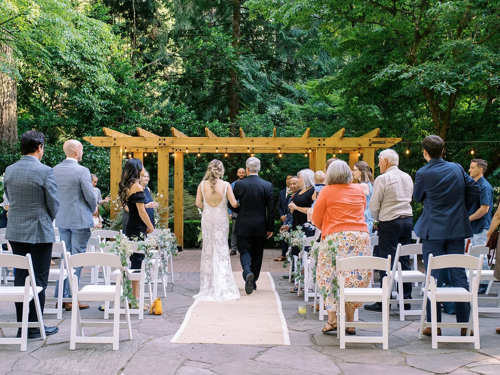 Bride walks down the garden aisle with a family member as guests stand for the ceremony.