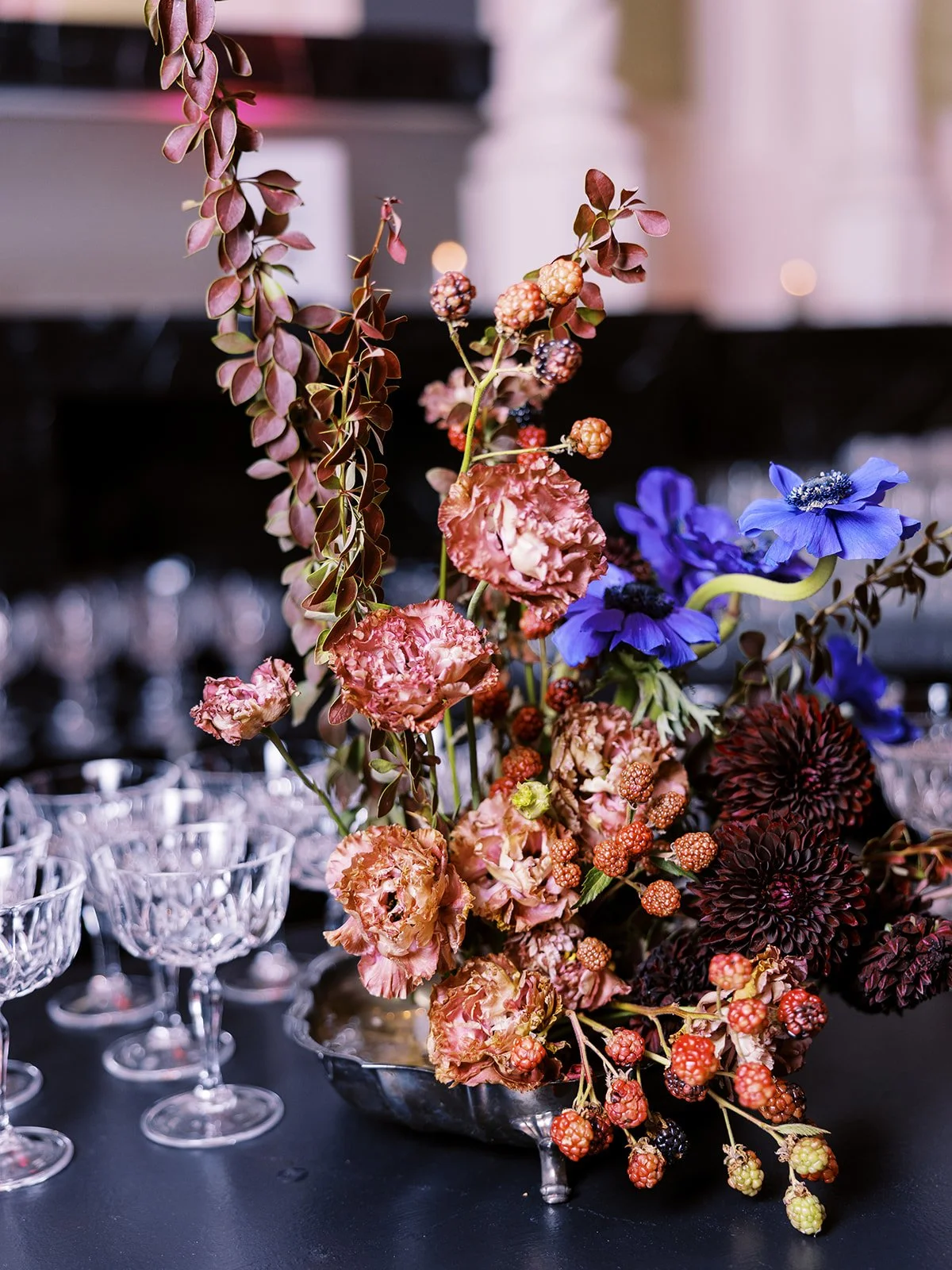 Colorful floral centerpiece with berries and textured blooms arranged beside rows of empty glassware.