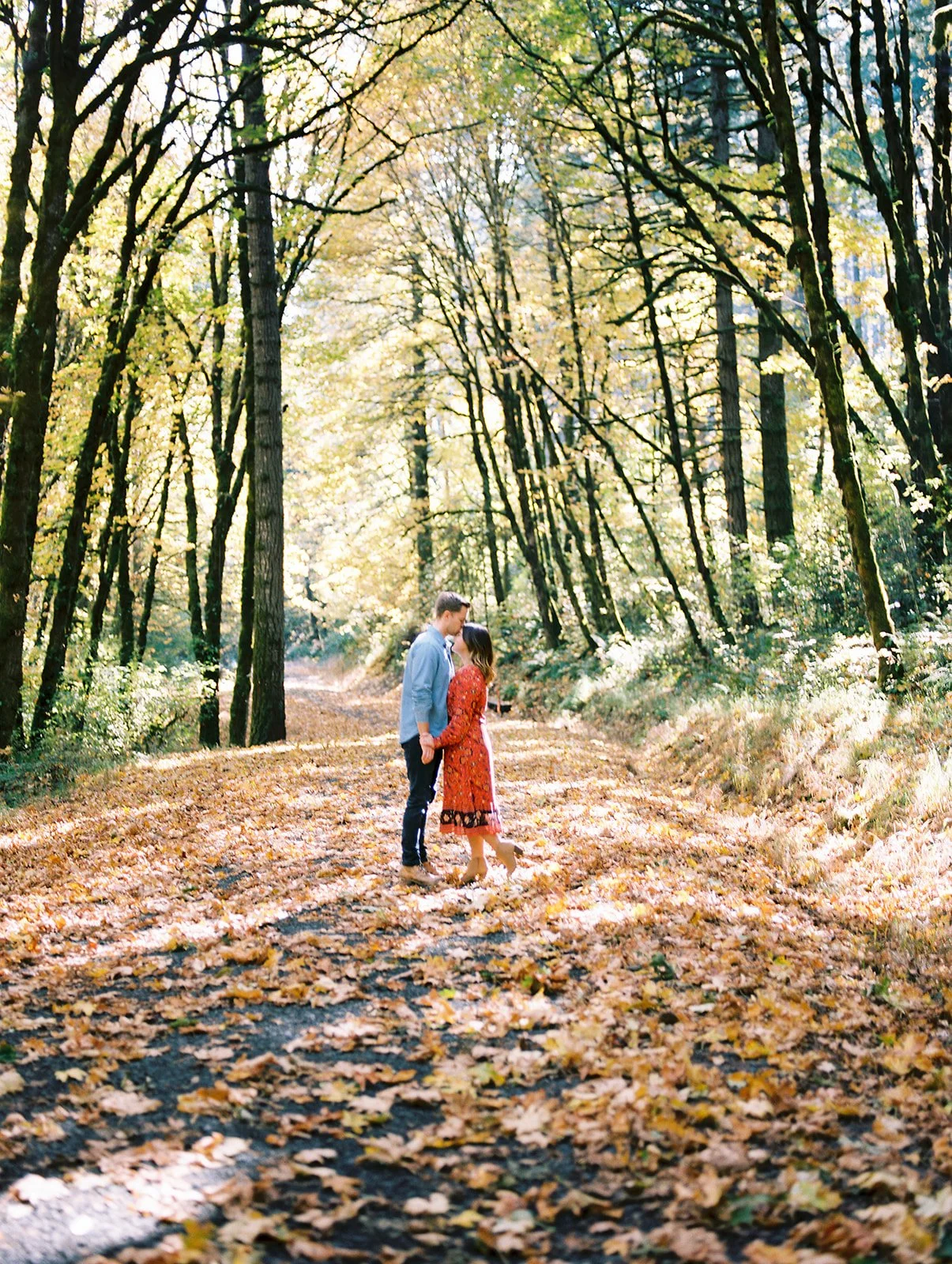 Couple standing on a leaf-covered forest path surrounded by tall trees during fall engagement photos Oregon