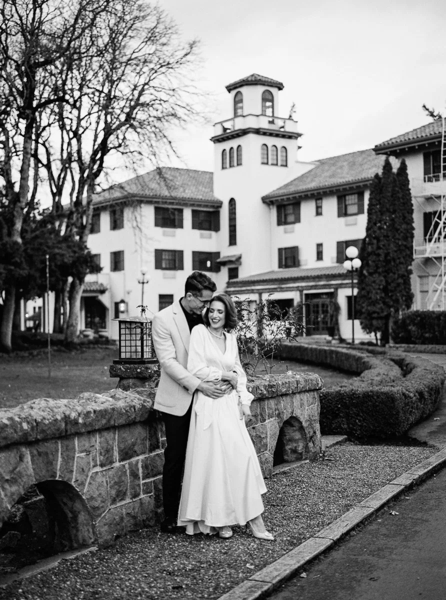 Black and white portrait of couple embracing by stone wall, woman in long dress smiling, man holding her from behind