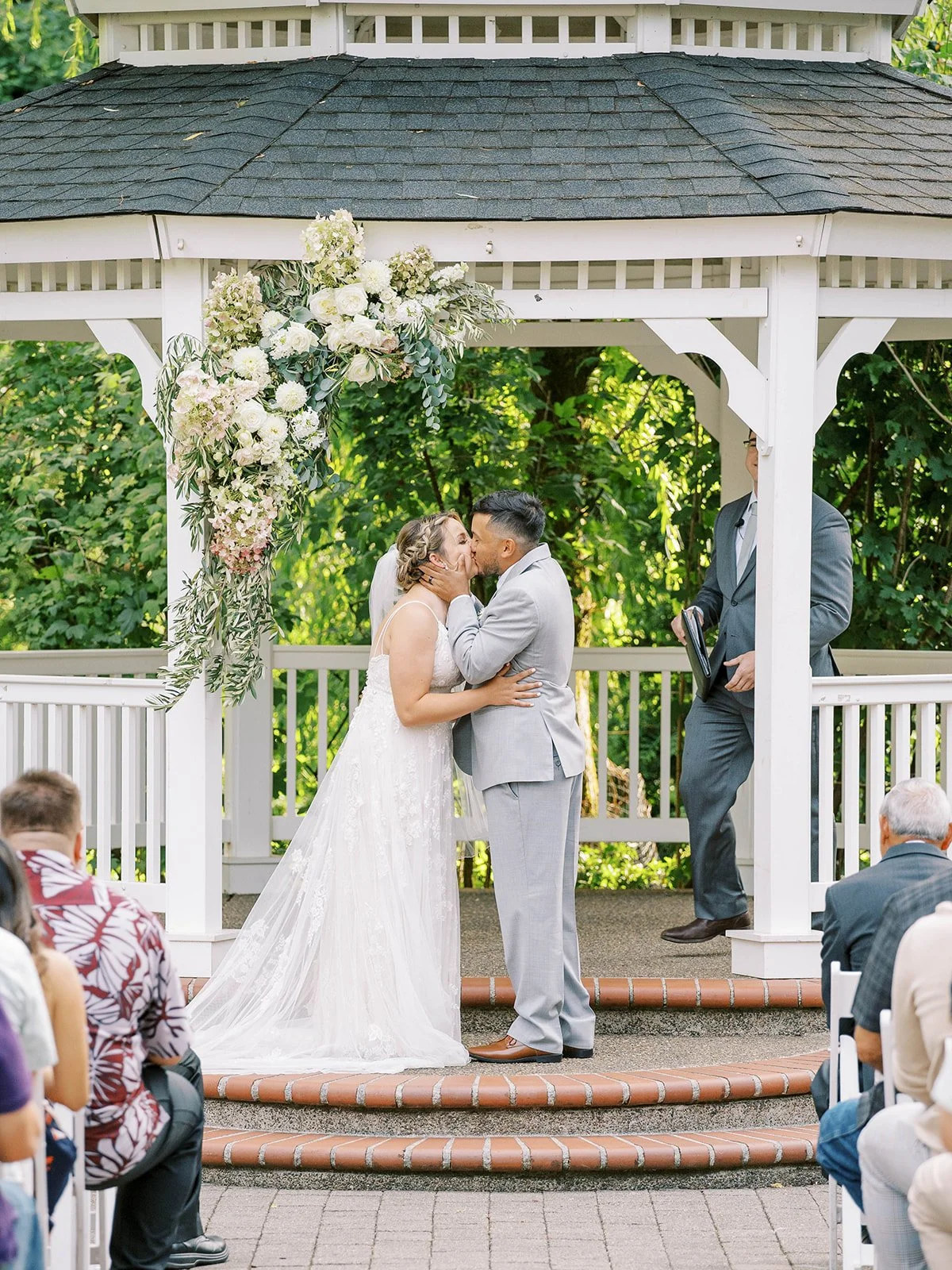 Bride and groom kiss beneath a white gazebo with cascading floral arrangement during an outdoor wedding ceremony, surrounded by seated guests.