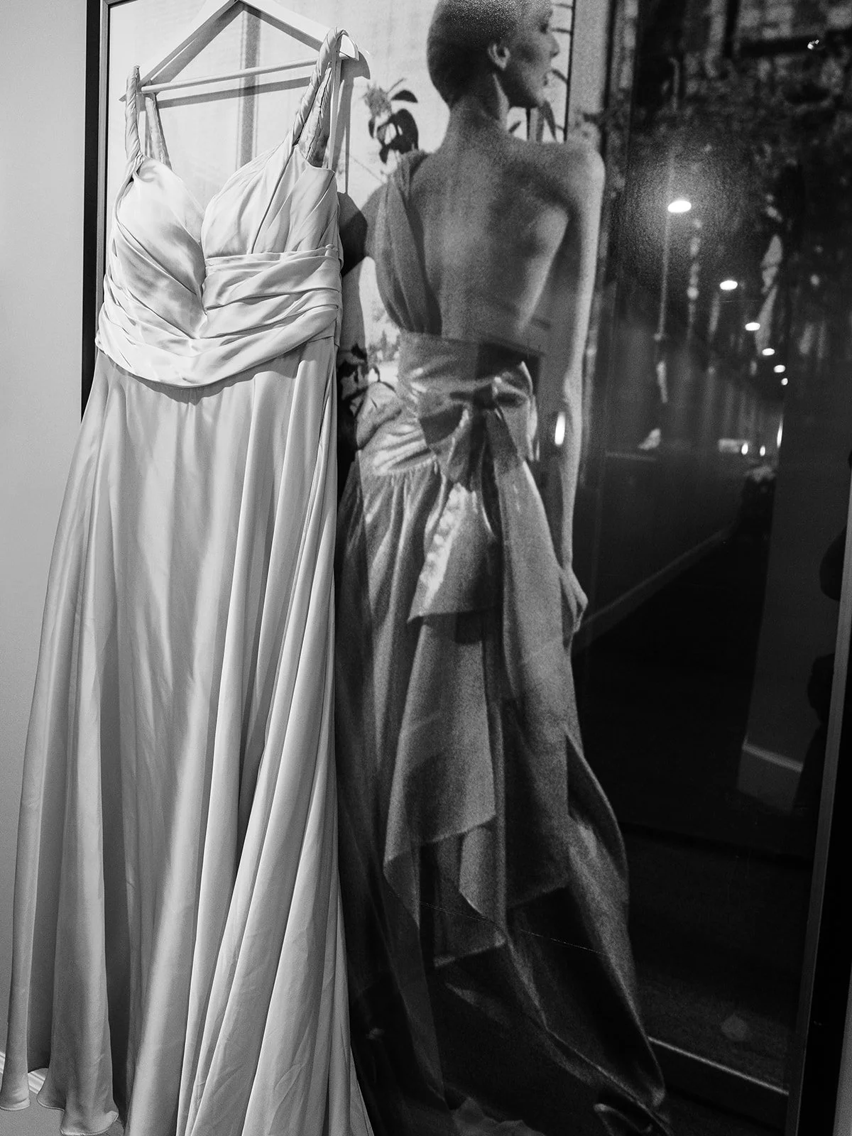 Silk wedding dress hanging on a rack beside a black-and-white fashion photograph.