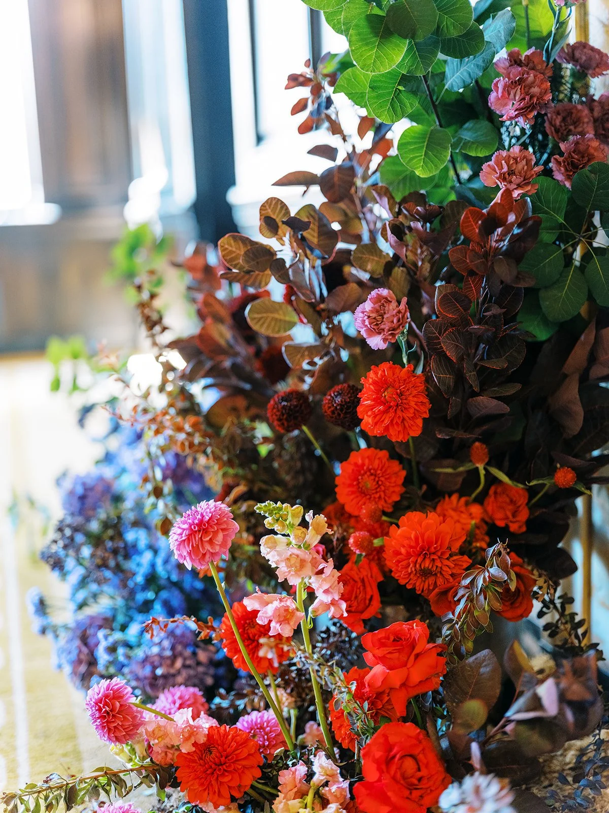 Colorful wedding floral installation with red, pink, and coral blooms arranged along a window-lit wall.