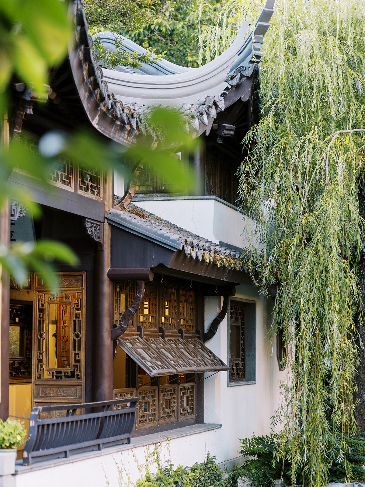 Architectural detail of Lan Su's multi-tiered roofline with sweeping upturned eaves, carved woodwork, latticed screens, and cascading willow branches.