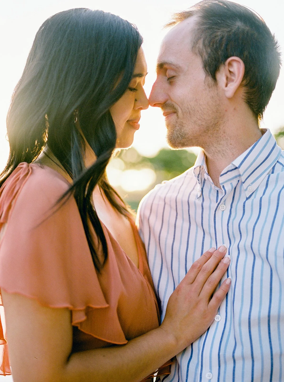 Close-up of couple touching foreheads at sunset, woman’s hand on partner’s striped shirt, warm golden light by the water