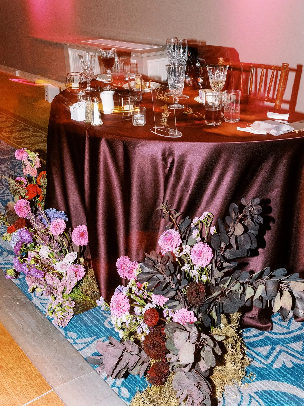 Reception table with burgundy linen, crystal glasses, and colorful floral arrangements along the floor.