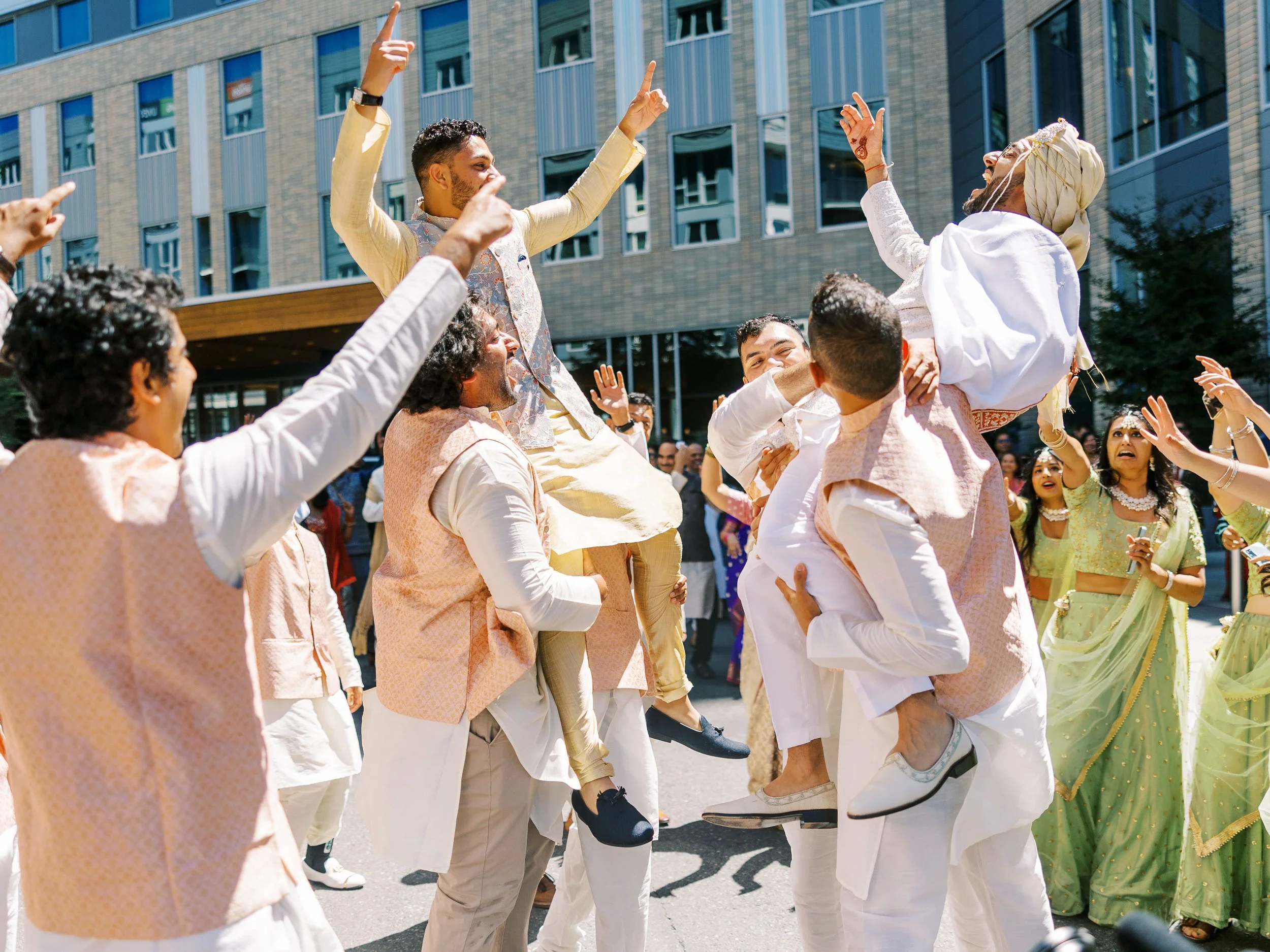 Wedding guests lift two men into the air during a lively outdoor Indian wedding celebration.
