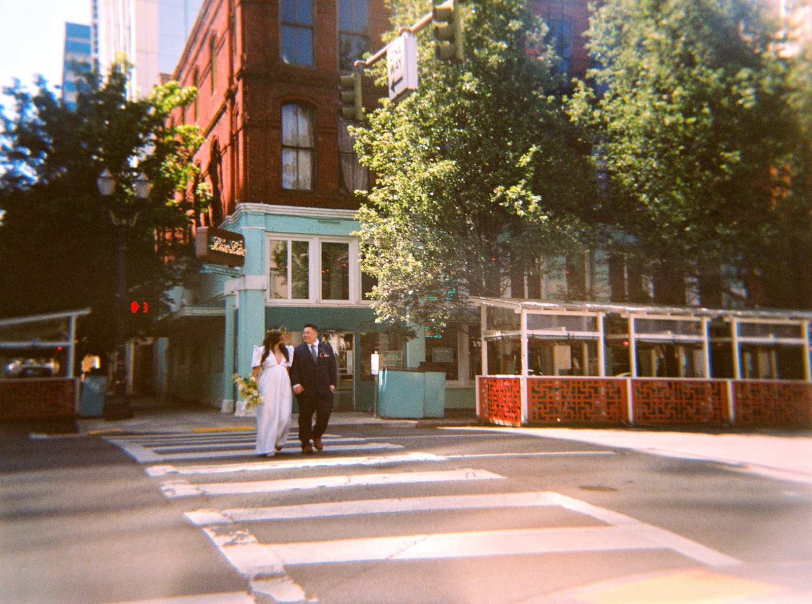 Newlyweds walk across a downtown Portland crosswalk, captured on film with colorful buildings and leafy trees behind them.