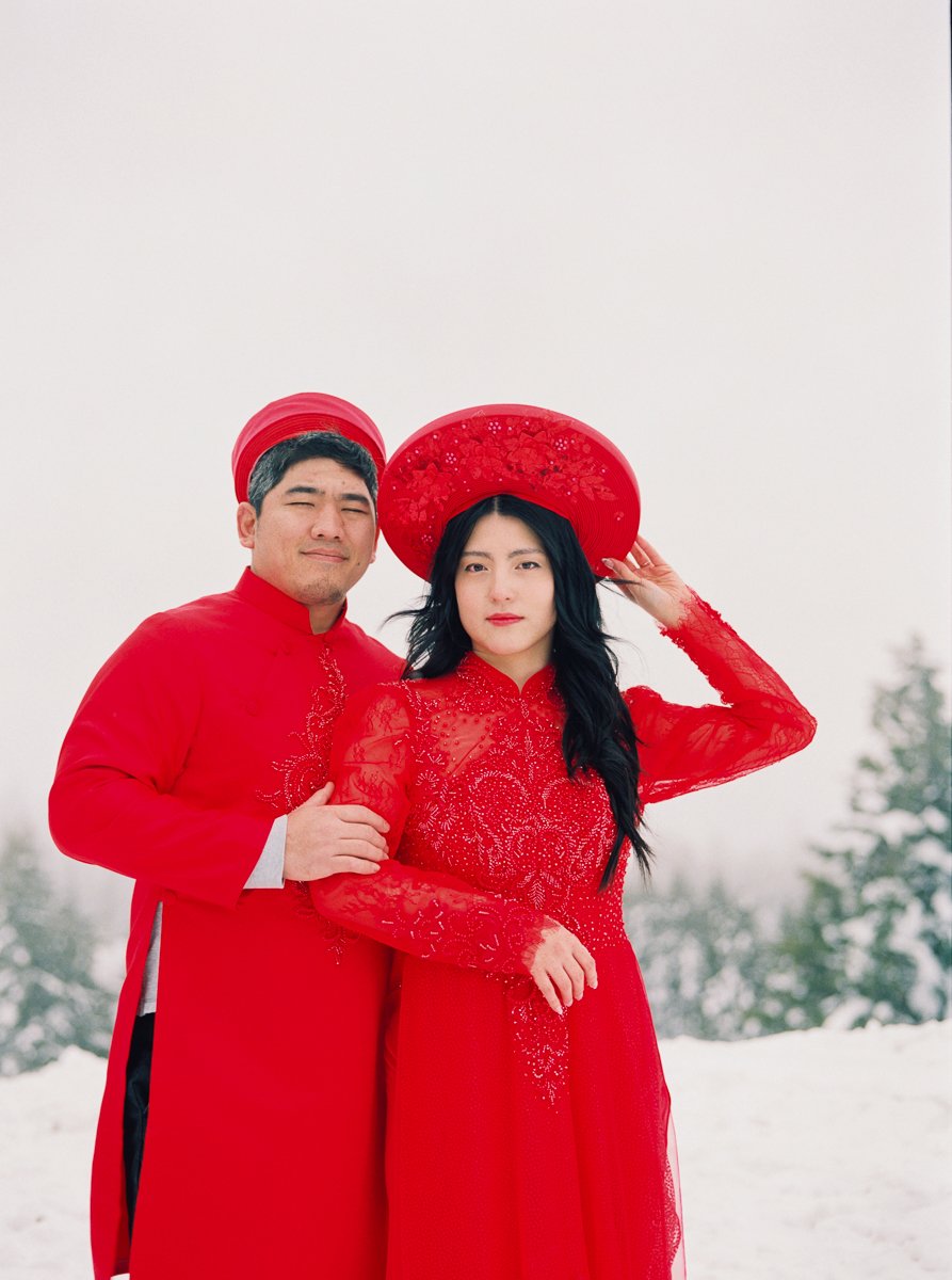 Couple in red Vietnamese áo dài posing together in snowy Mount Hood setting.