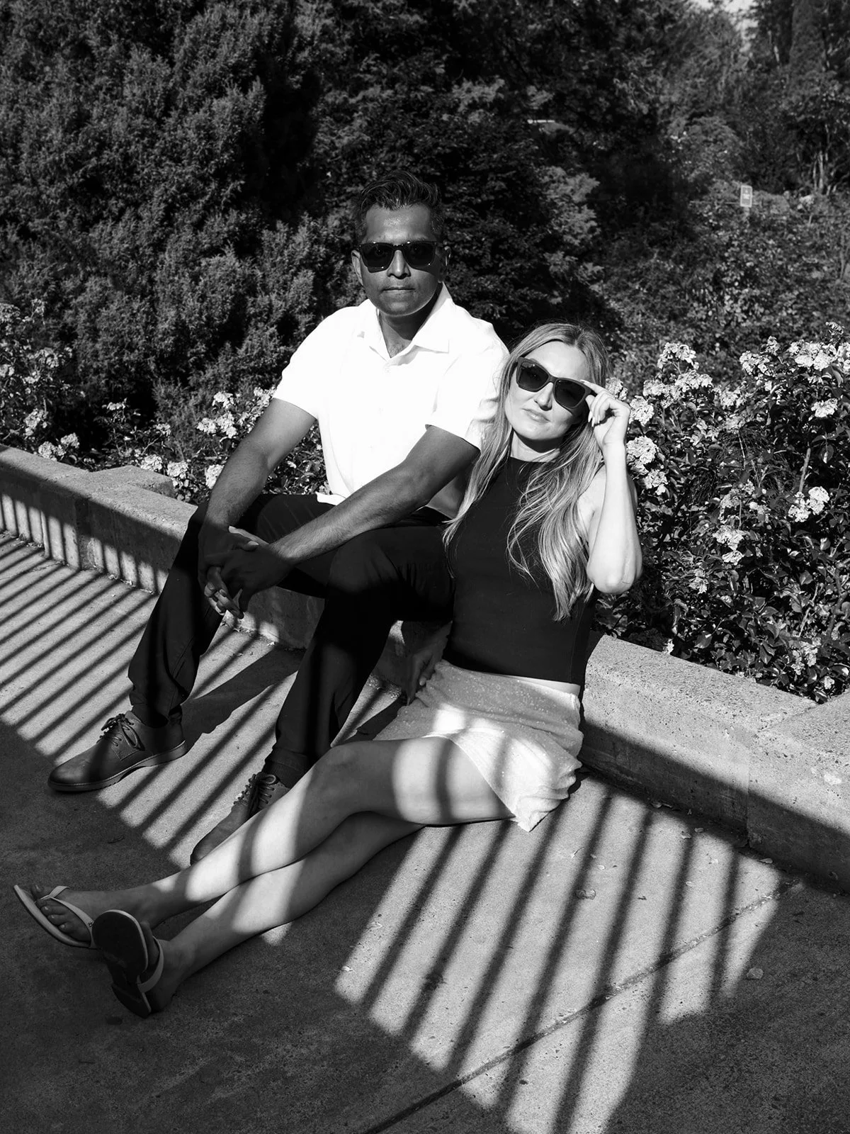 Black and white portrait of a couple in sunglasses lounging on a garden wall in dramatic striped shadows.