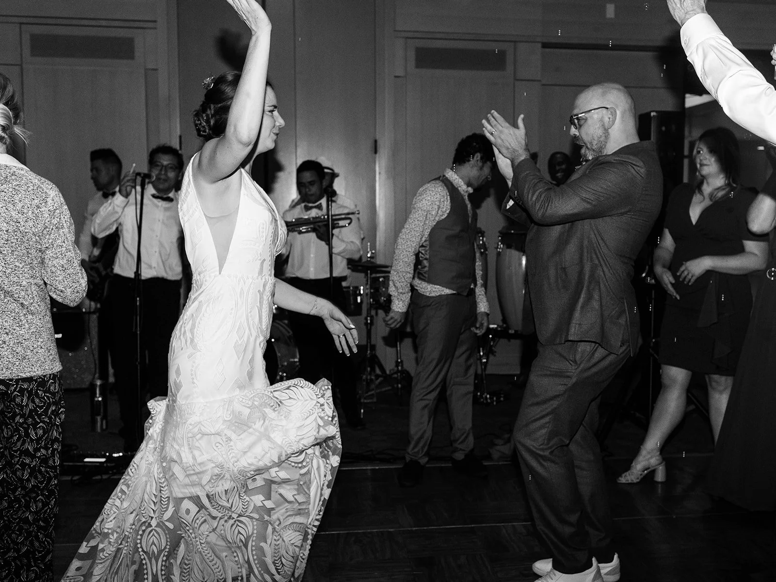 Black and white photo of the bride twirling on the dance floor with her arm raised, groom clapping nearby and the band behind them