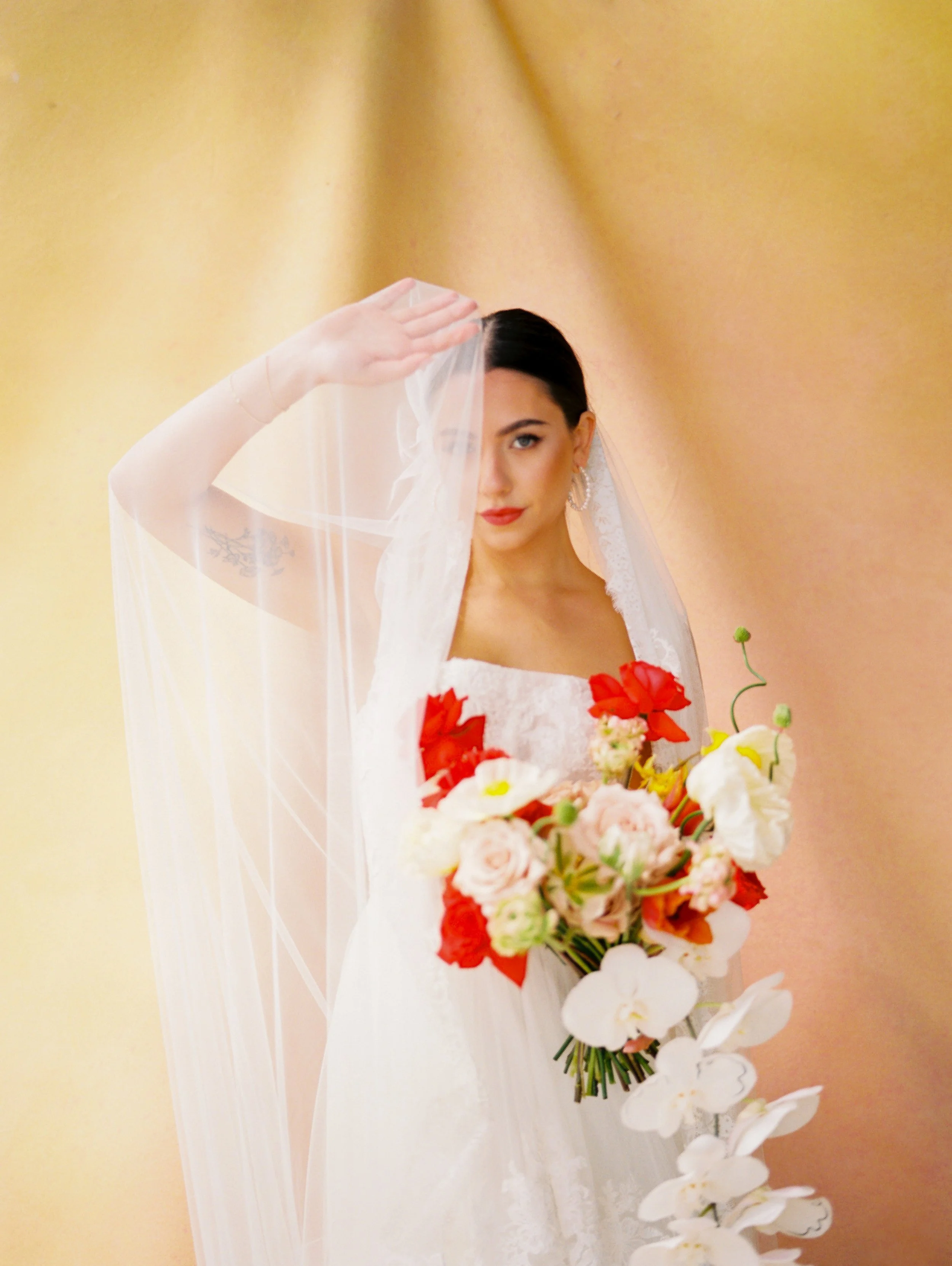 Bride in a lace wedding gown holding a red, white and blush bouquet beneath a sheer lace veil.