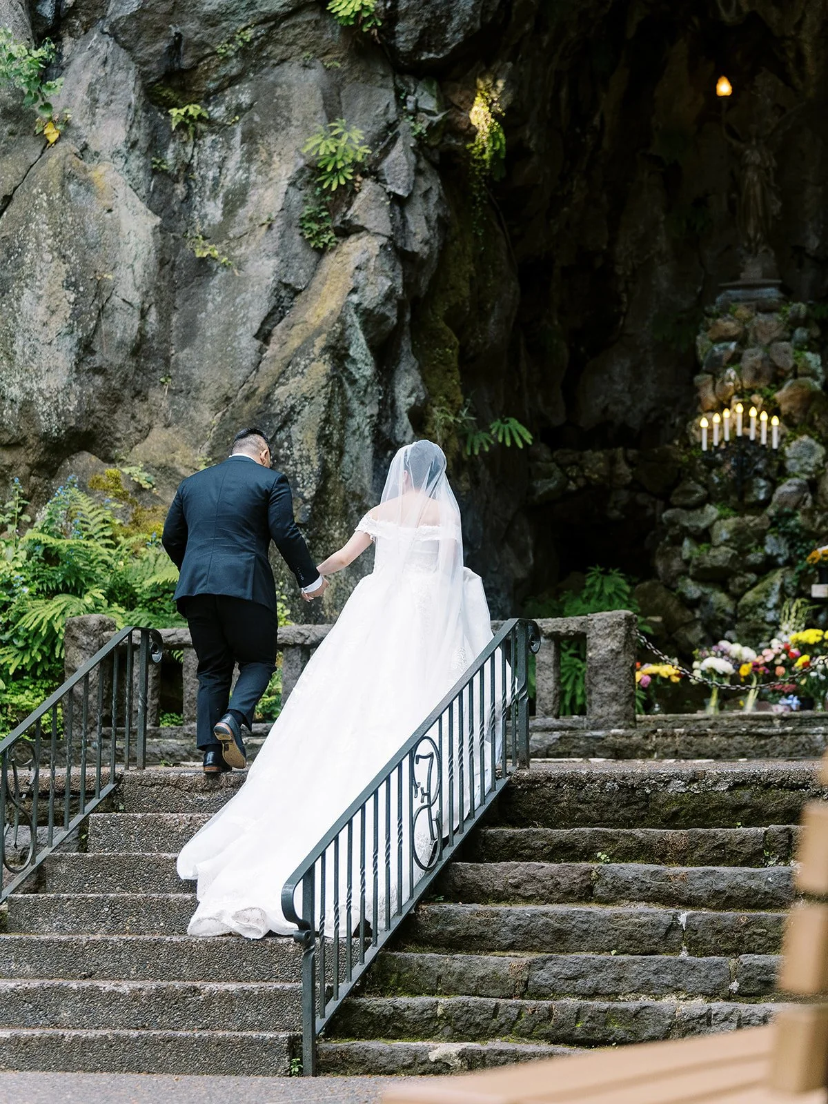 Bride and groom seen from behind climbing the mossy stone steps toward The Grotto's cave altar, candles glowing inside the shrine