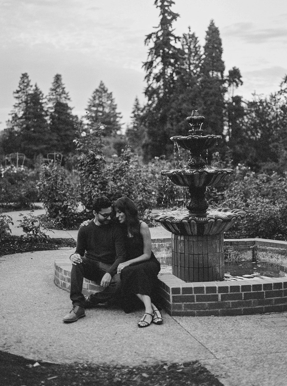 Black-and-white portrait of a couple sitting closely beside a fountain in the rose garden.