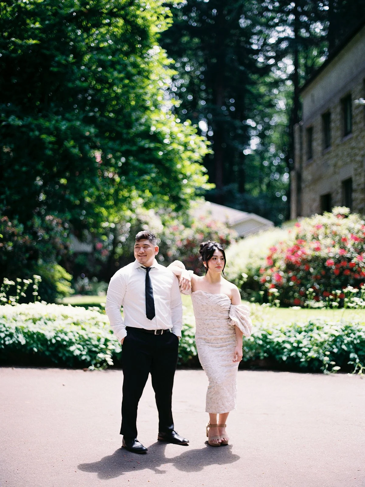 Bride resting her arm on the groom's shoulder as they pose on a sun-dappled path at The Grotto, red and pink rhododendrons in the background