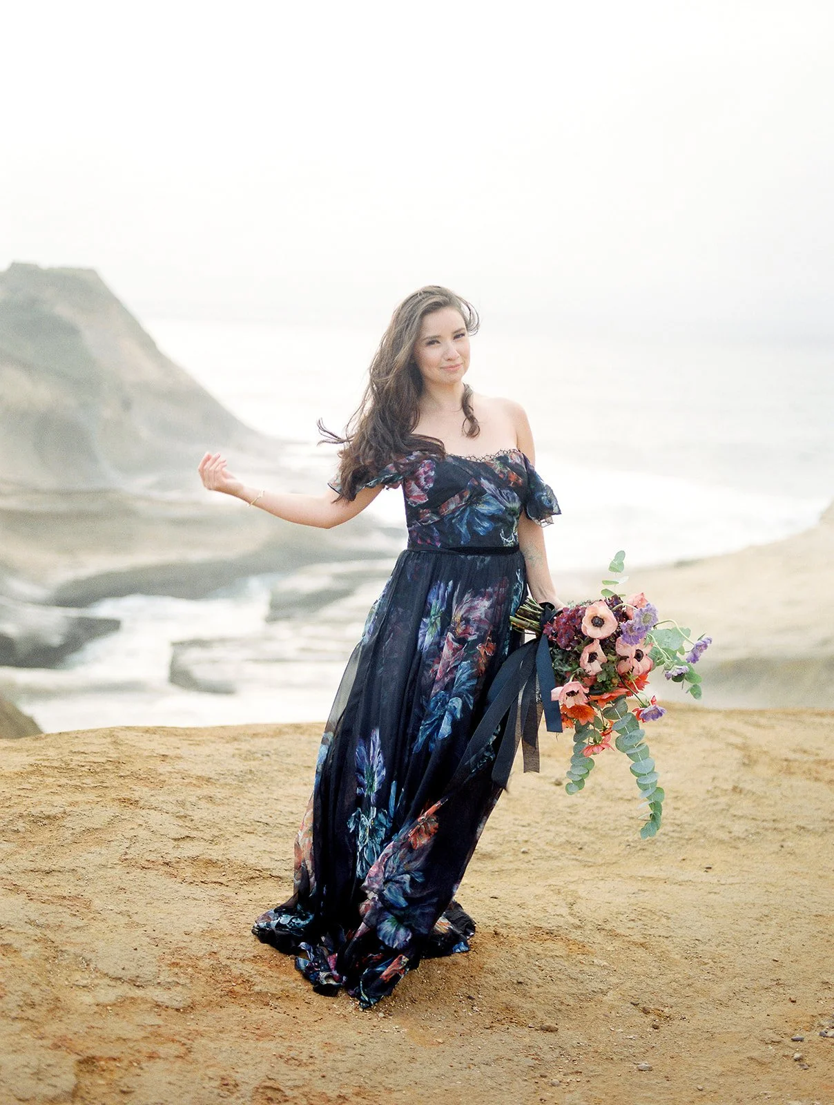 Woman in flowing floral dress holding bouquet on seaside cliff with ocean and rocky shoreline behind